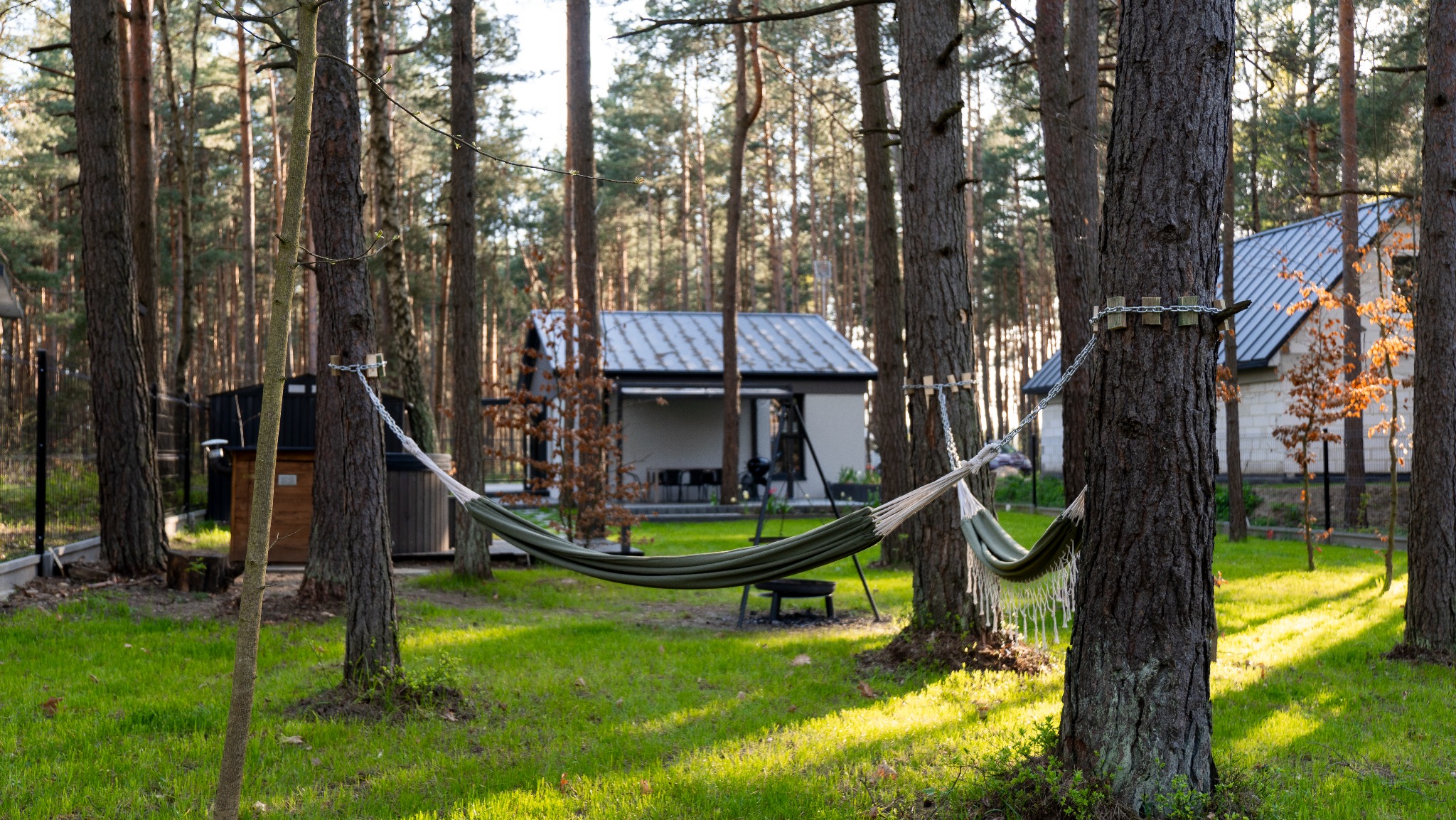 Leśny krajobraz z hamakiem zawieszonym między drzewami, w tle domki letniskowe z szarymi dachami, zielona trawa i drzewa iglaste.