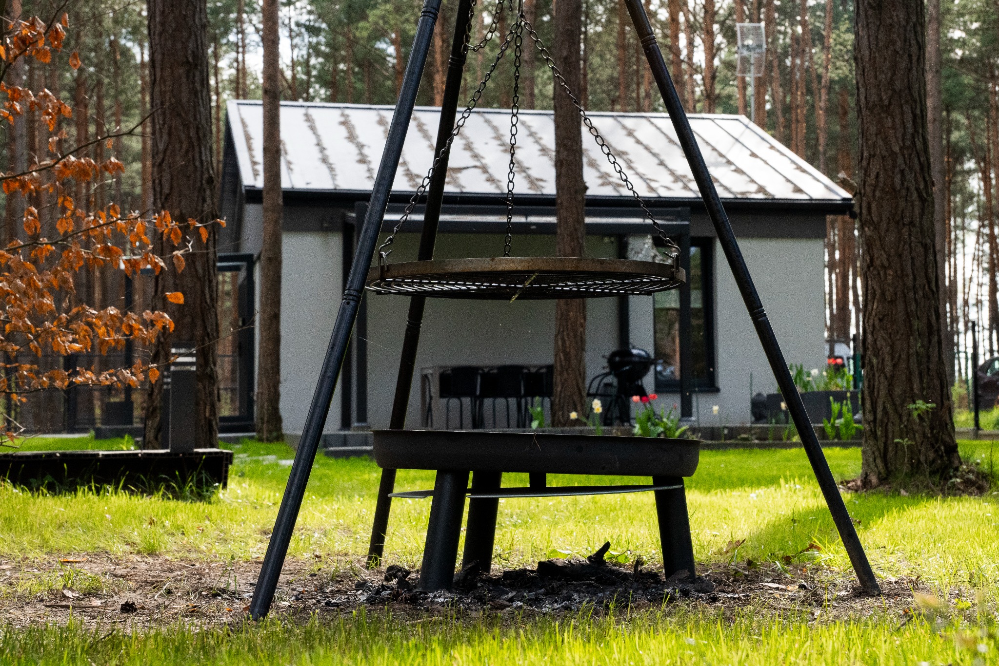 Ognisko ogrodowe z rusztem i paleniskiem na tle leśnego domku z białymi ścianami i ciemną stolarką. Ujęcie w słoneczny dzień, wśród drzew i zielonej trawy.