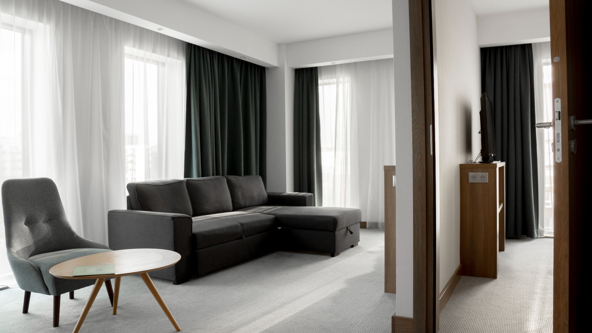 Nowoczesny pokój hotelowy z szarym narożnikiem, fotelem i stolikiem kawowym. W tle okna z zasłonami w odcieniach szarości i bieli. Widok na telewizor przez otwarte drzwi.