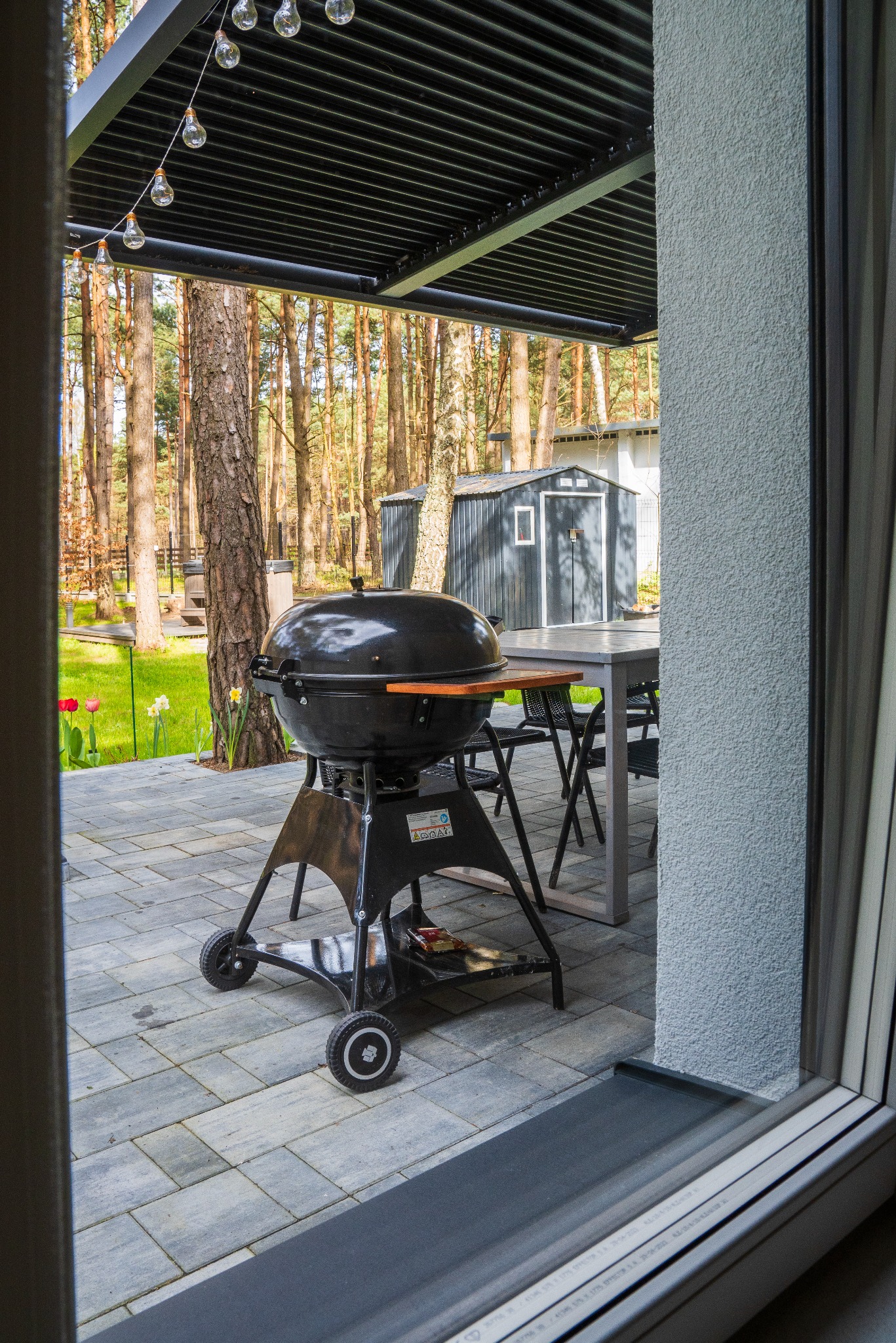 Czarny grill na kółkach na szarym patio, widok zza okna. W tle las i altana, oświetlona girlandą żarówek. Wiosenne kwiaty na trawniku dodają koloru.