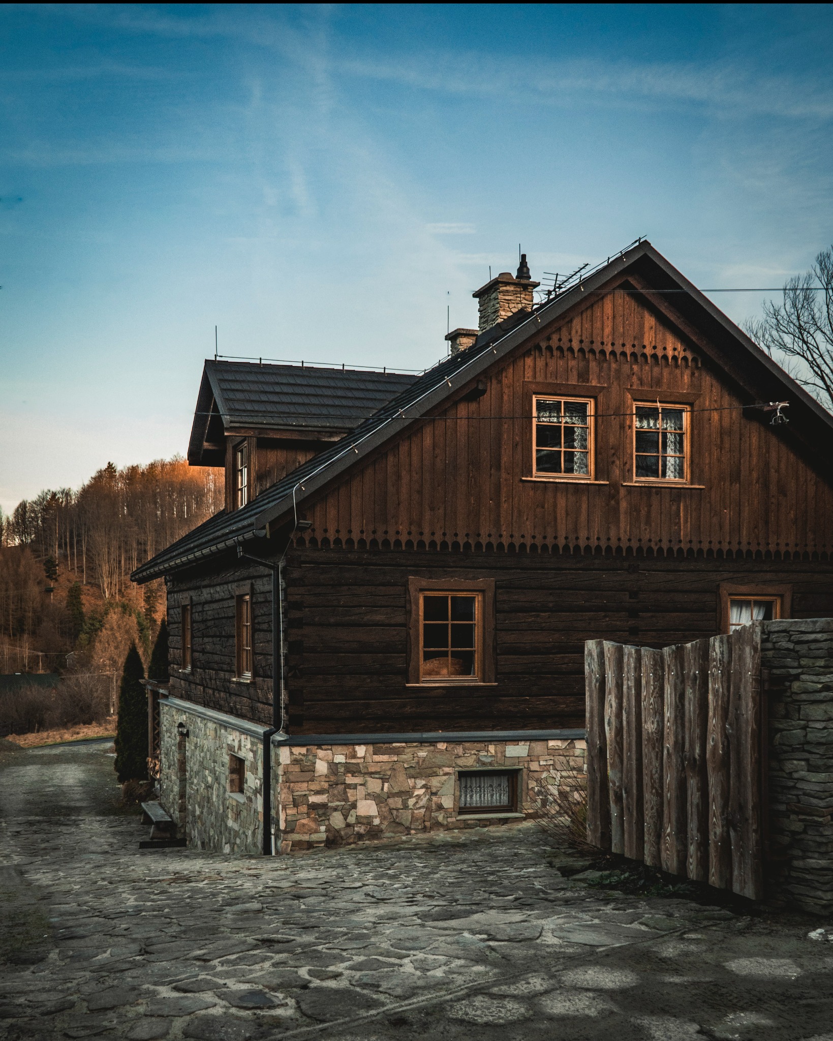 Drewniany dom w góralskim stylu z kamienną podmurówką i brukowaną drogą. Widoczne okna z drewnianymi ramami i zdobienia. Tło stanowią drzewa i błękitne niebo.