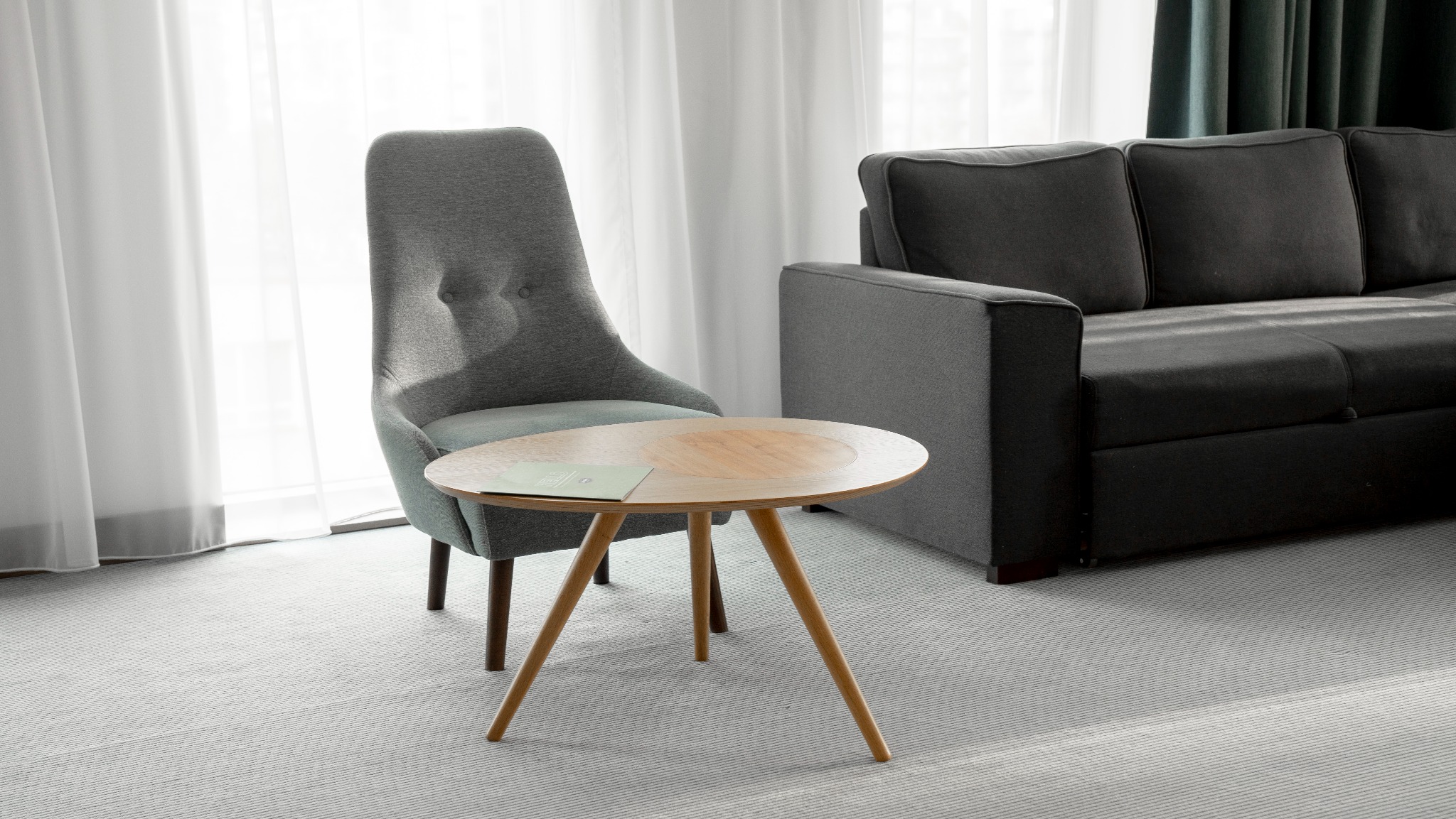 Jasne wnętrze: szary fotel, drewniany stolik z ulotką, fragment szarej sofy, białe zasłony. Minimalistyczny design, stonowana kolorystyka, spokojna atmosfera.
