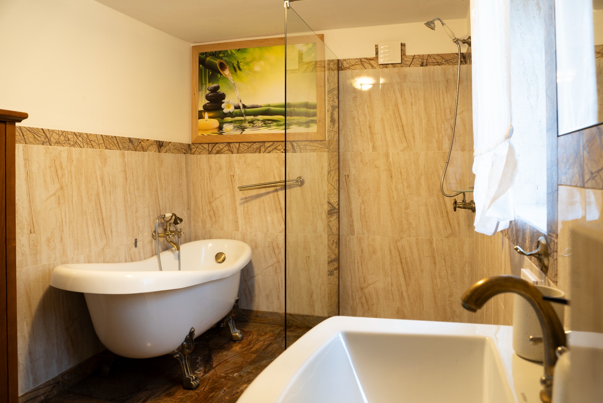 Elegancka łazienka z wanną na lwich nóżkach, beżowymi płytkami i szklanym panelem prysznicowym. Na ścianie obrazek z motywem zen.
