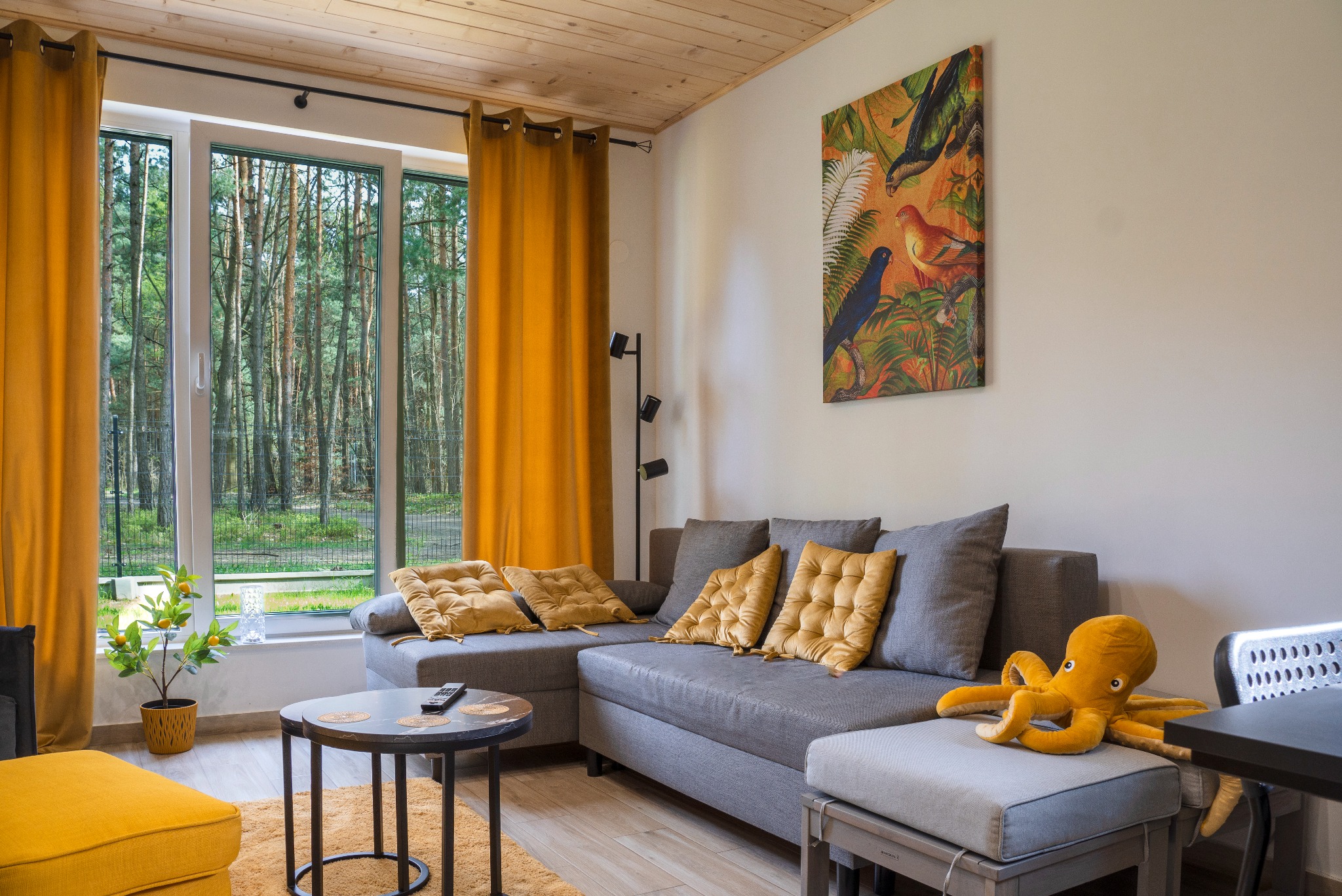 Jasne wnętrze salonu z szarą sofą, żółtymi akcentami (poduszki, zasłony), widokiem na las i obrazem z motywem ptaków. Pluszowy ośmiornica na pufie.