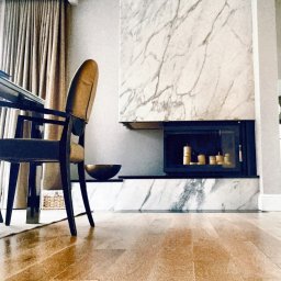 Granmarys S C M.Siuda R.Siuda - Marmurowy kominek z czarnym wkładem, dekoracyjne świece w środku, parkiet na podłodze. Widok z niskiego kąta, z perspektywą na krzesło i stół w tle.