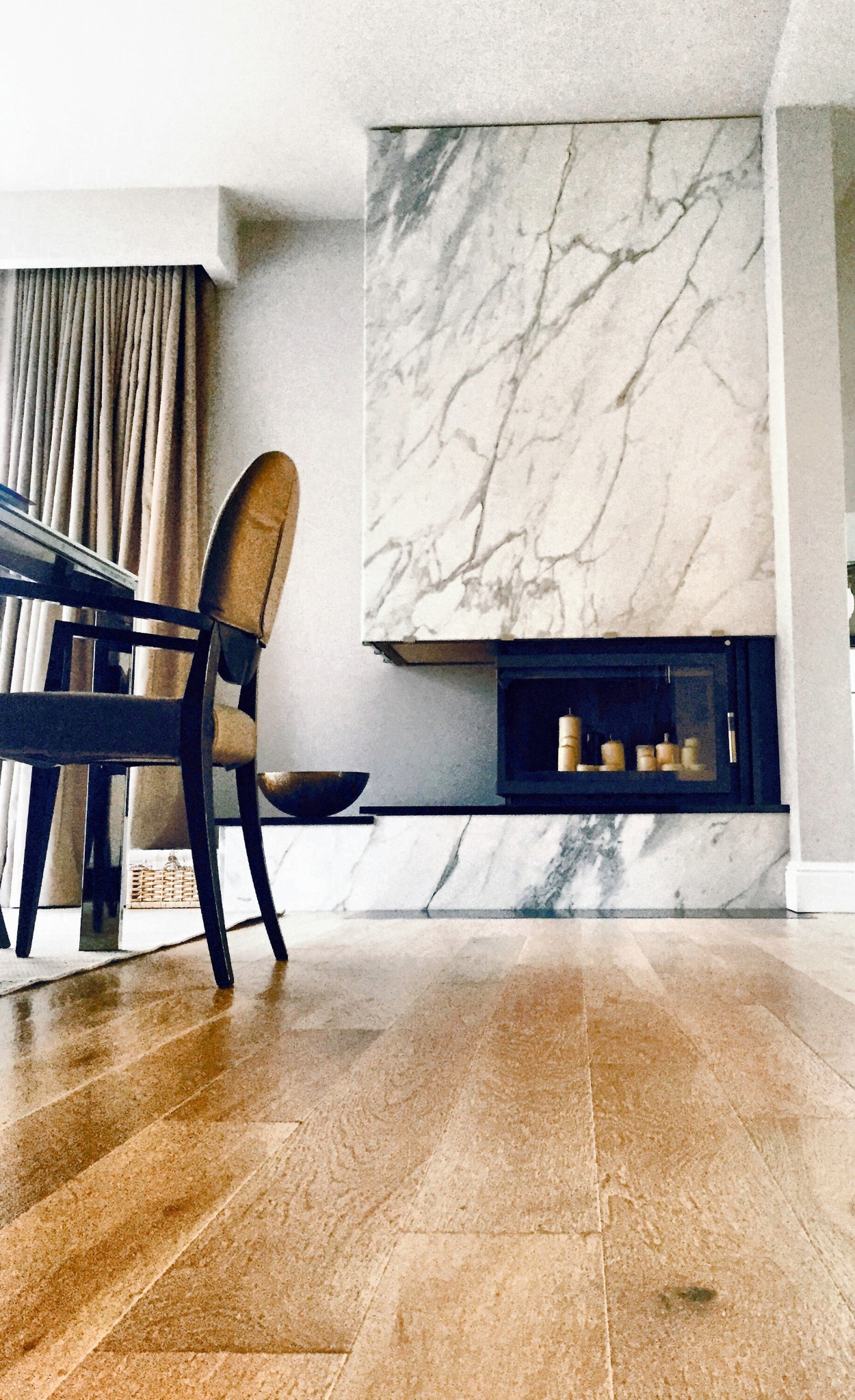 Marmurowy kominek z czarnym wkładem, dekoracyjne świece w środku, parkiet na podłodze. Widok z niskiego kąta, z perspektywą na krzesło i stół w tle.