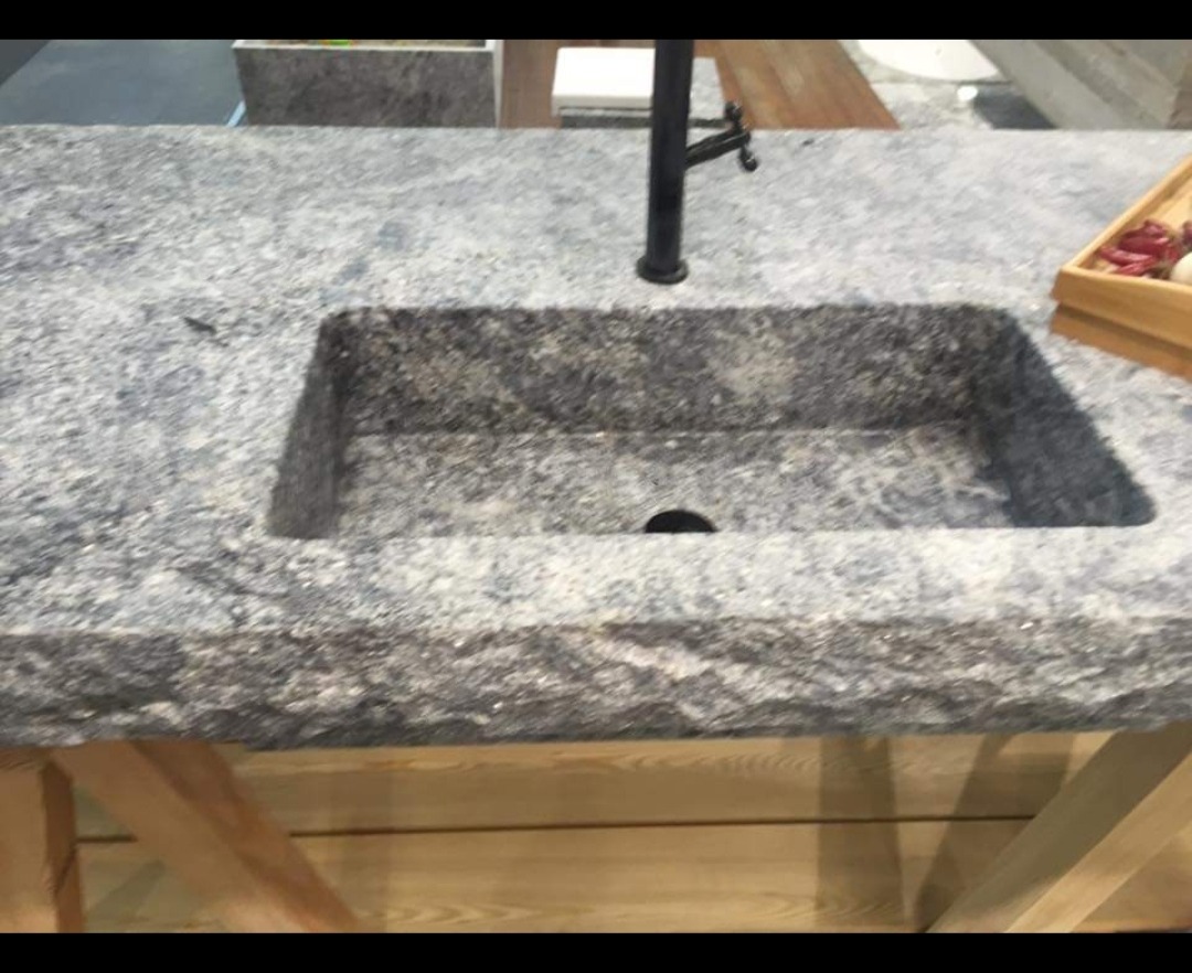 Granitowy blat kuchenny z wbudowanym zlewem o surowym wykończeniu i czarną armaturą, osadzony na drewnianej konstrukcji. Widoczny fragment drewnianej tacy.