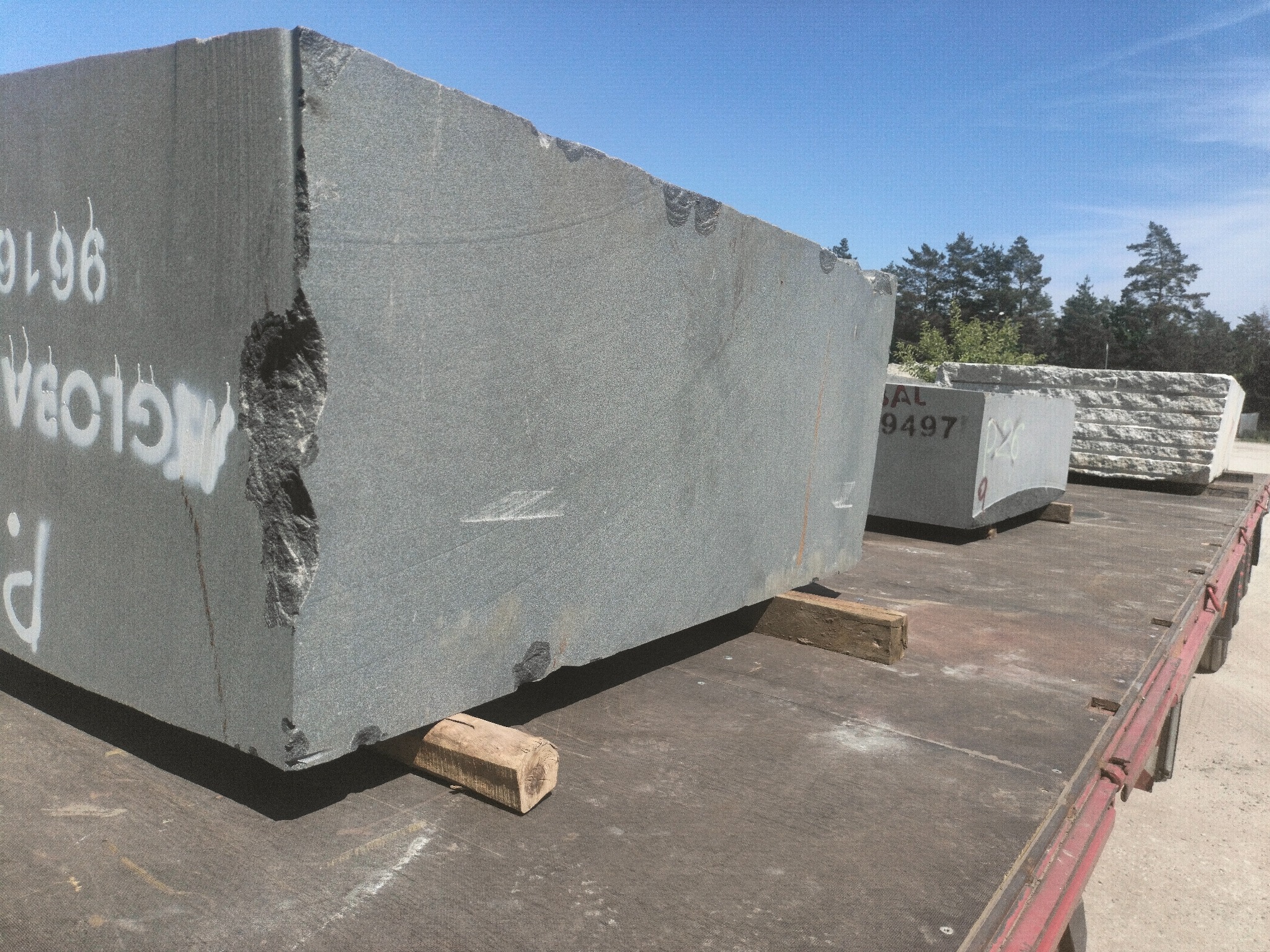 Surowe bloki szarego granitu na platformie transportowej, z widocznymi oznaczeniami i uszkodzeniami krawędzi, na tle błękitnego nieba i drzew.