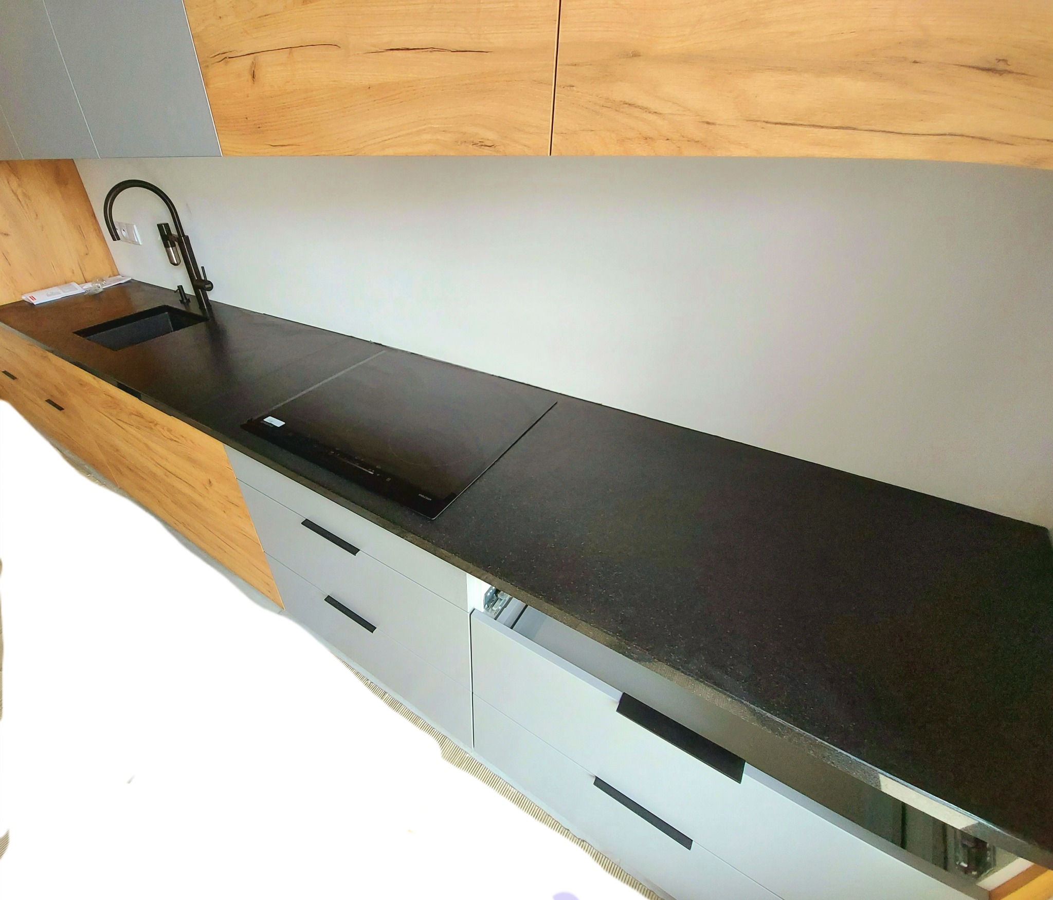 Czarny blat granitowy w kuchni z płytą indukcyjną i minimalistycznym zlewem, satynowe wykończenie, jasne drewniane i szare szafki, nowoczesny design.