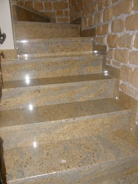 Kamienne schody wewnętrzne z beżowego granitu, dopasowane do kamiennej ściany, z delikatnym oświetleniem punktowym na krawędziach stopni.