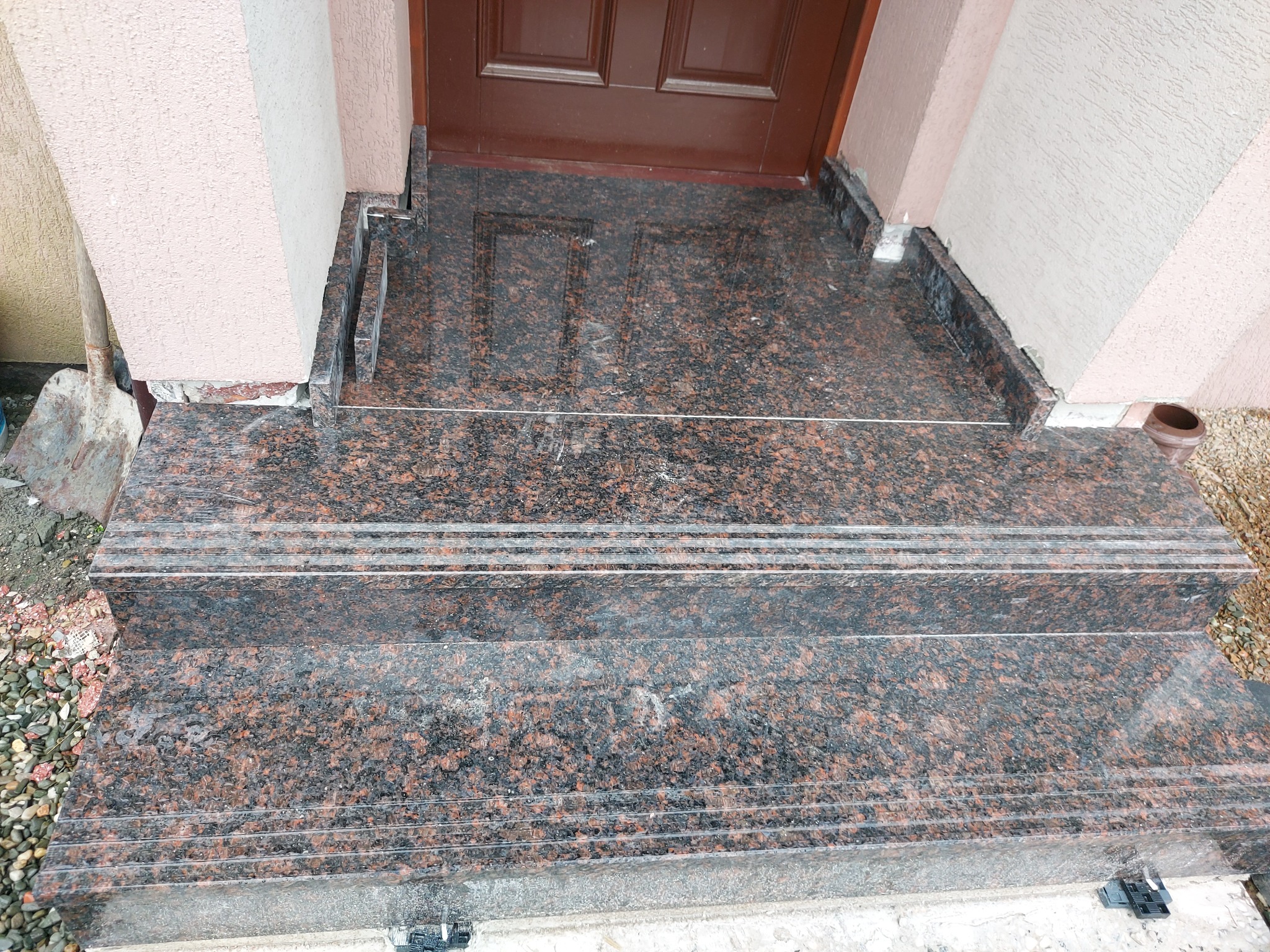 Granitowe schody zewnętrzne prowadzące do brązowych drzwi wejściowych. Widoczna łopata oparta o ścianę. Detal wykończenia z brązowego kamienia.