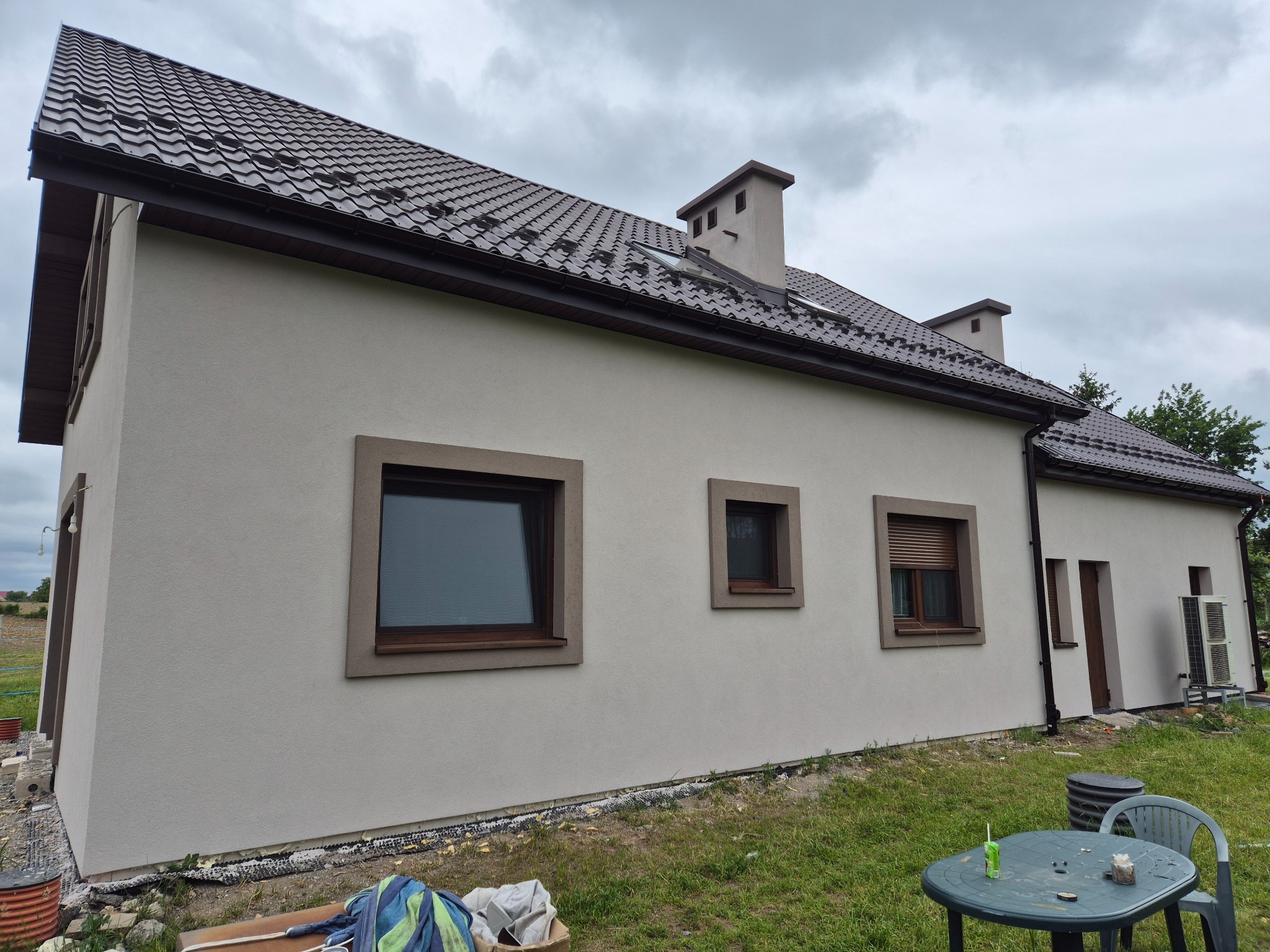 Jasna elewacja domu jednorodzinnego z brązowymi obramowaniami okien i dachu, widok z boku, na trawniku stolik i krzesła.