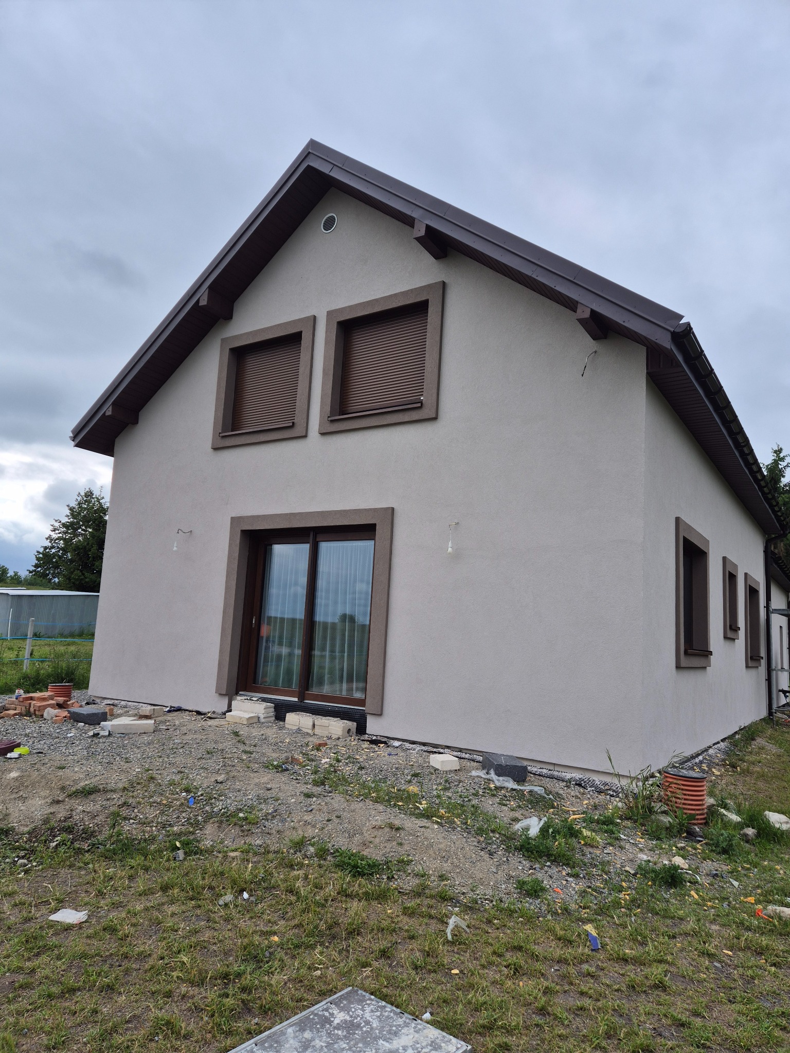 Dom jednorodzinny z beżową elewacją i brązowymi ramami okiennymi, widoczny z perspektywy żabiej, na tle pochmurnego nieba i zielonego trawnika z gruzem.