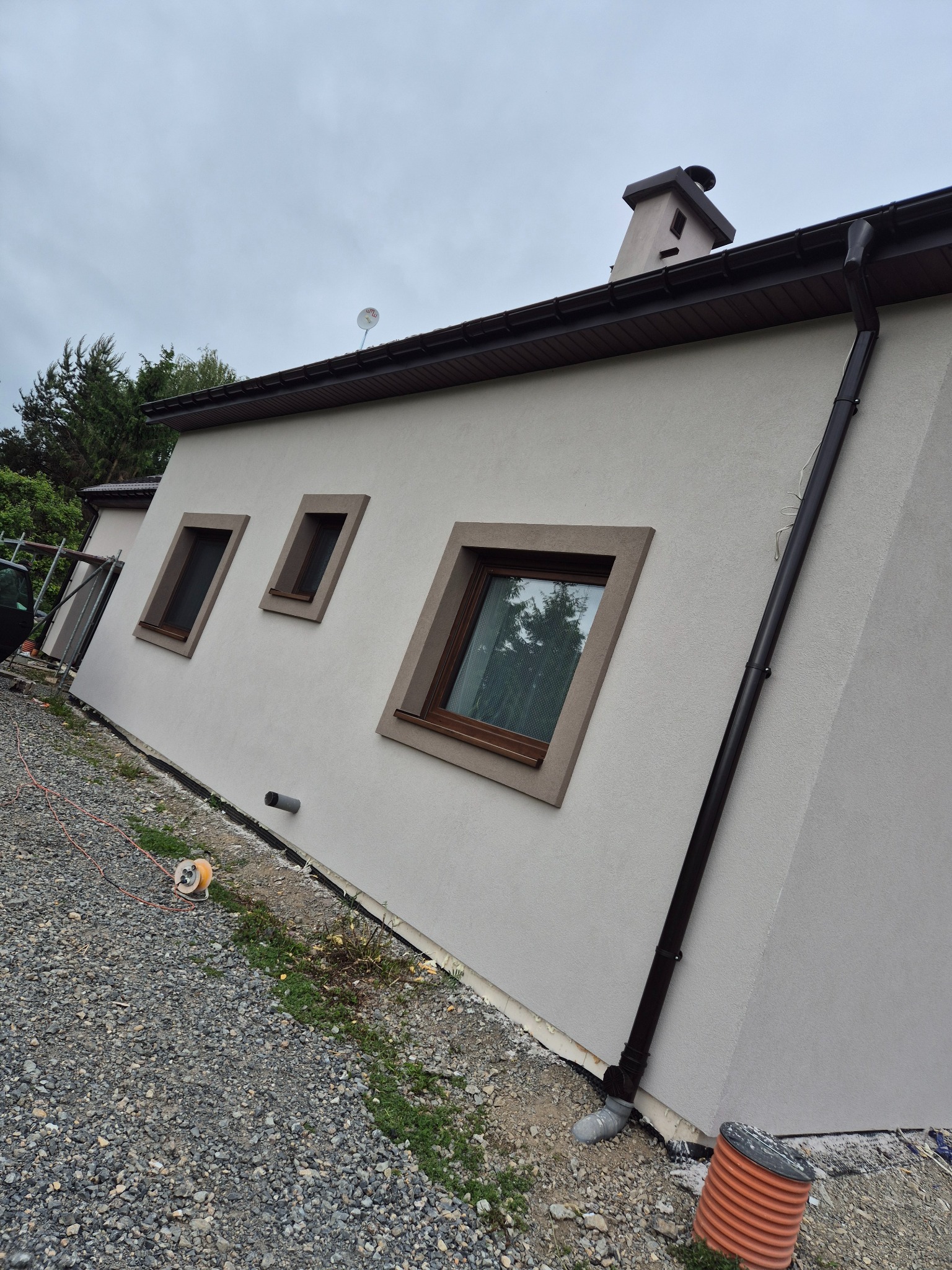 Świeżo otynkowana elewacja domu jednorodzinnego z brązowymi ramami okiennymi i rynną. Widok z boku na szarą fasadę, z drobnym żwirkiem na ziemi.