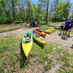 Spływy kajakowe Eskulapa - Grupa osób przygotowuje się do spływu kajakowego; widoczne kolorowe kajaki na brzegu rzeki, zalesione otoczenie i osoby w kajakach na wodzie w tle.