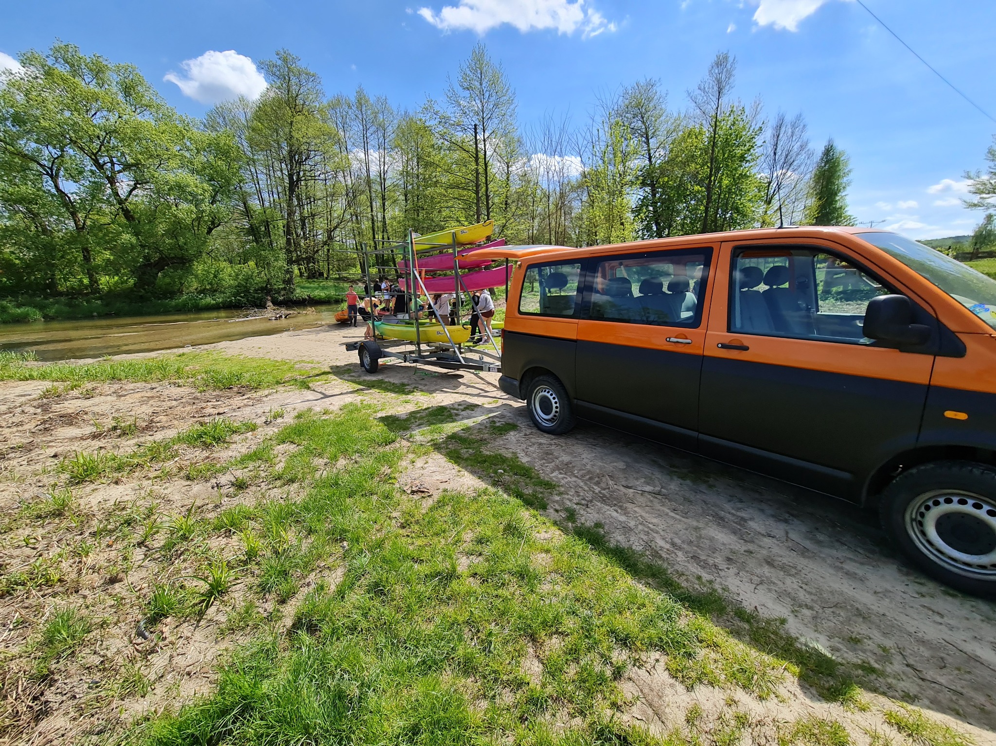 Pomarańczowo-czarny bus z przyczepą pełną kajaków nad brzegiem rzeki, przygotowania do spływu kajakowego w słoneczny dzień. Grupa osób w tle.