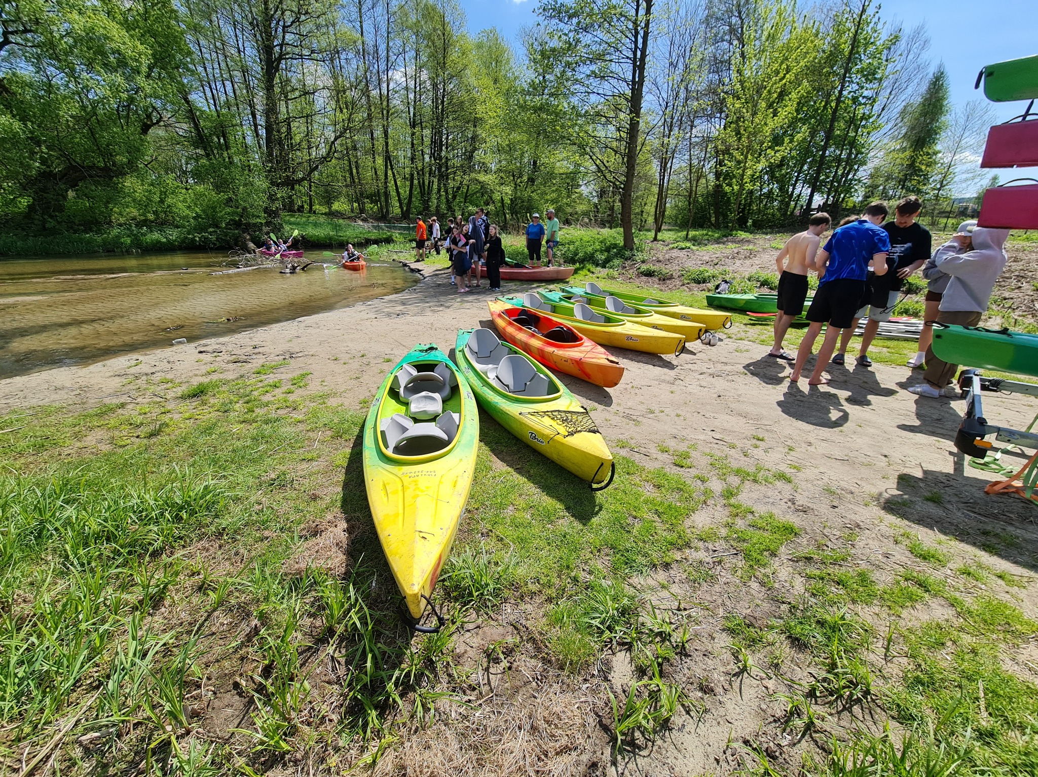 Grupa osób przygotowuje się do spływu kajakowego; widoczne kolorowe kajaki na brzegu rzeki, zalesione otoczenie i osoby w kajakach na wodzie w tle.