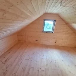 Drewniane wnętrze poddasza z oknem dachowym. Widoczne deski na podłodze, ścianach i suficie, tworzące ciepły, naturalny klimat. Surowe wykończenie.