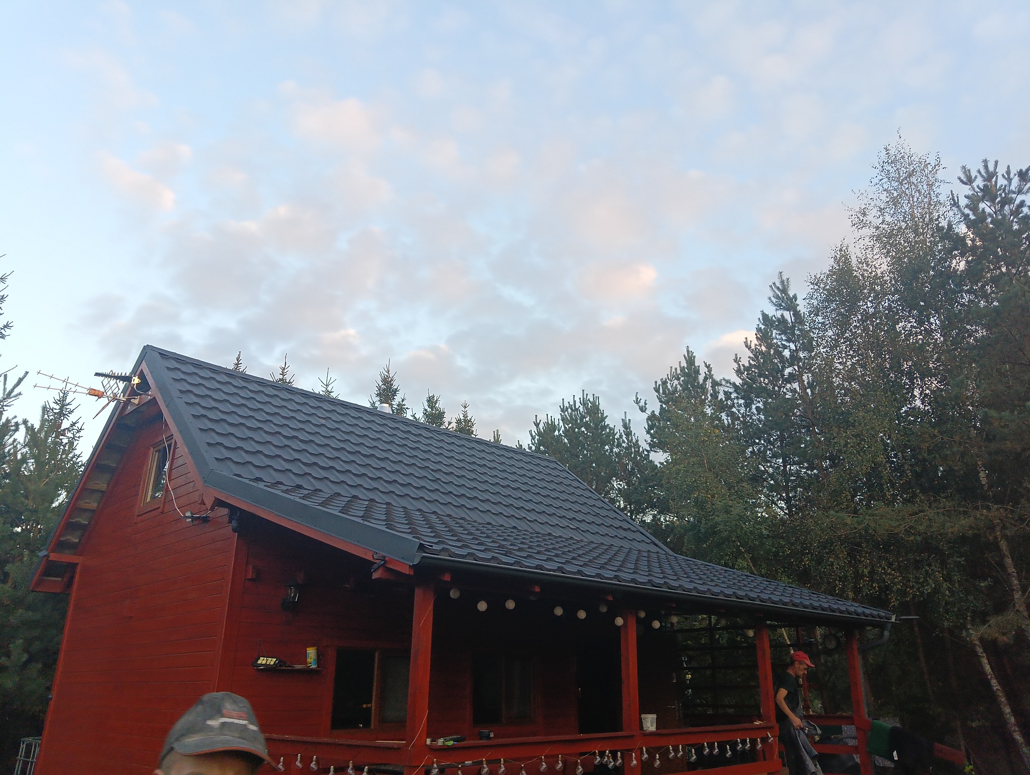 Czerwony dom z bali z czarnym dachem na tle drzew i pochmurnego nieba. Widoczne elementy dekoracyjne na werandzie i antena na dachu.