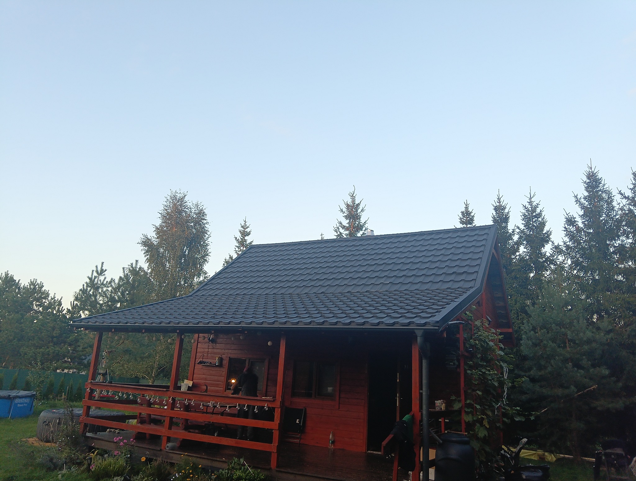 Czerwony dom z bali z zadaszonym tarasem i ciemnym dachem na tle drzew iglastych i błękitnego nieba. Widok z dołu, w tle basen Bestway.