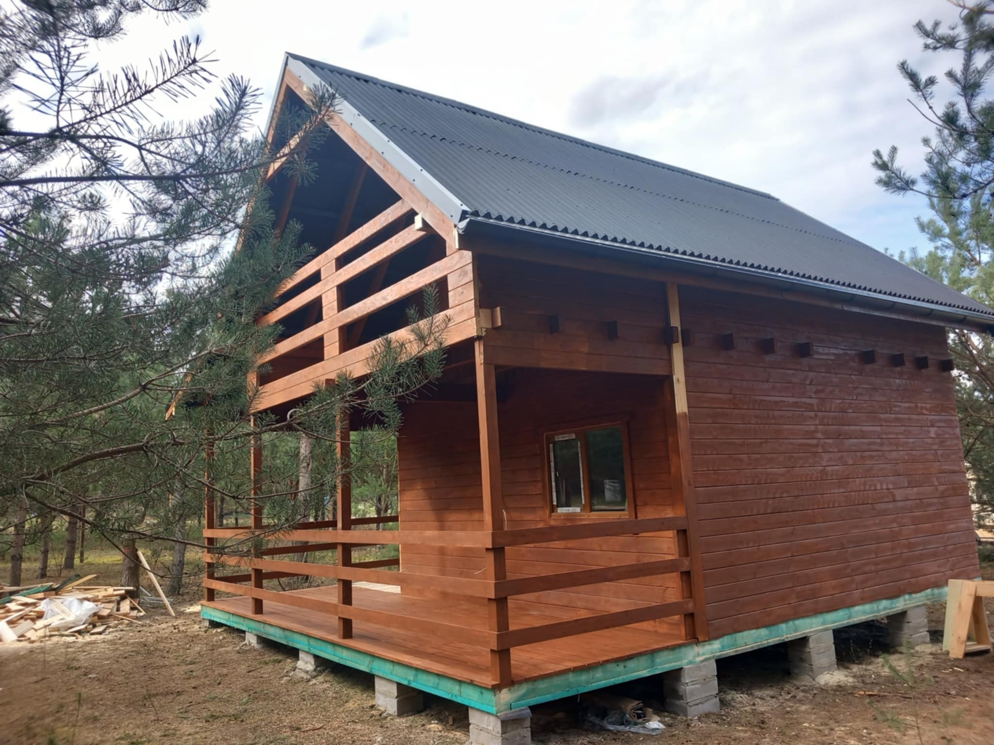 Drewniany domek letniskowy z zadaszonym tarasem w Smętówko. Widoczne bale pomalowane na brązowo, szary dach i betonowe bloczki fundamentowe.