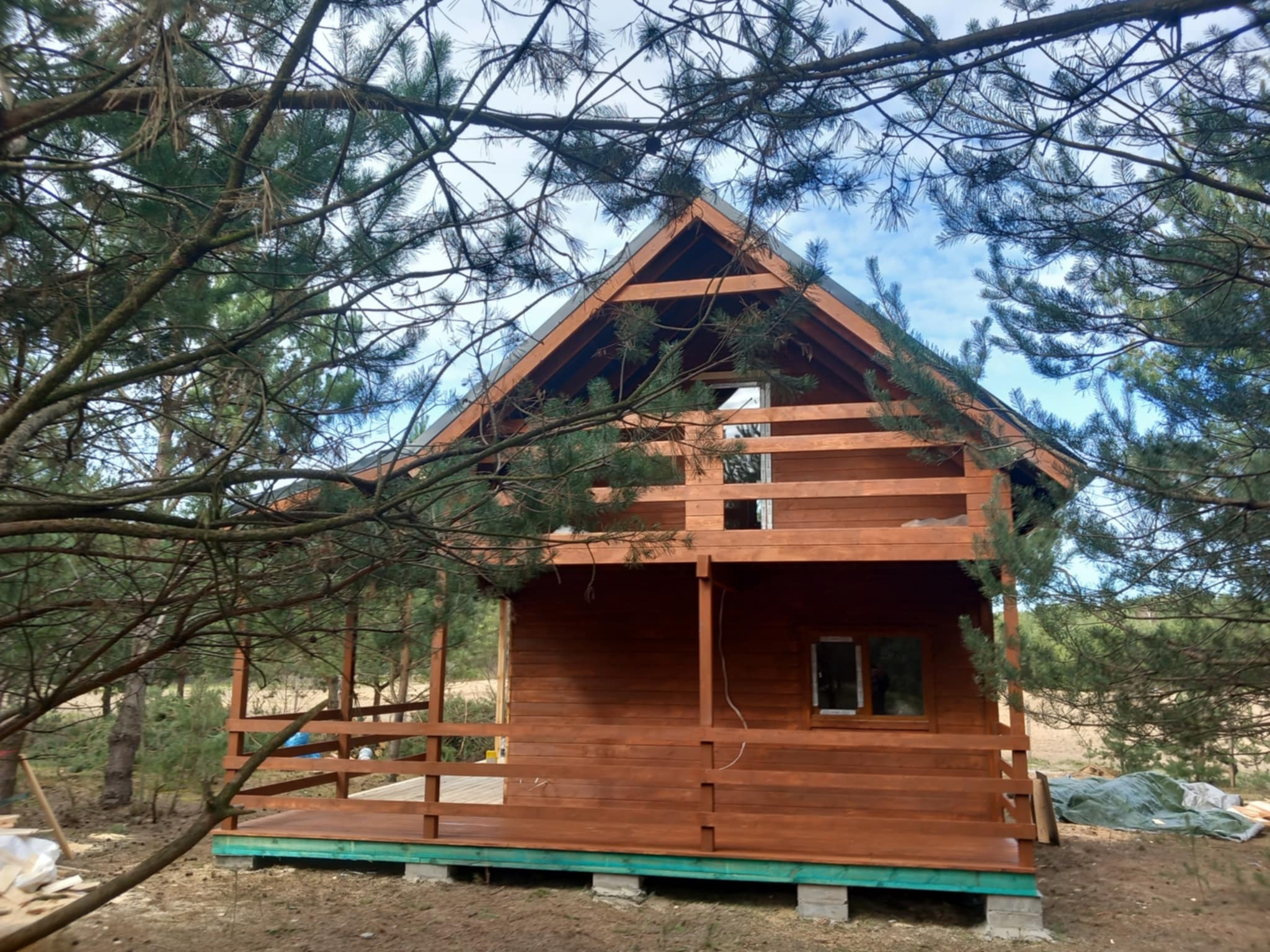 Brązowy, drewniany domek letniskowy z tarasem, widziany przez gałęzie drzew. Dom na betonowych bloczkach, z oknami i dwuspadowym dachem.