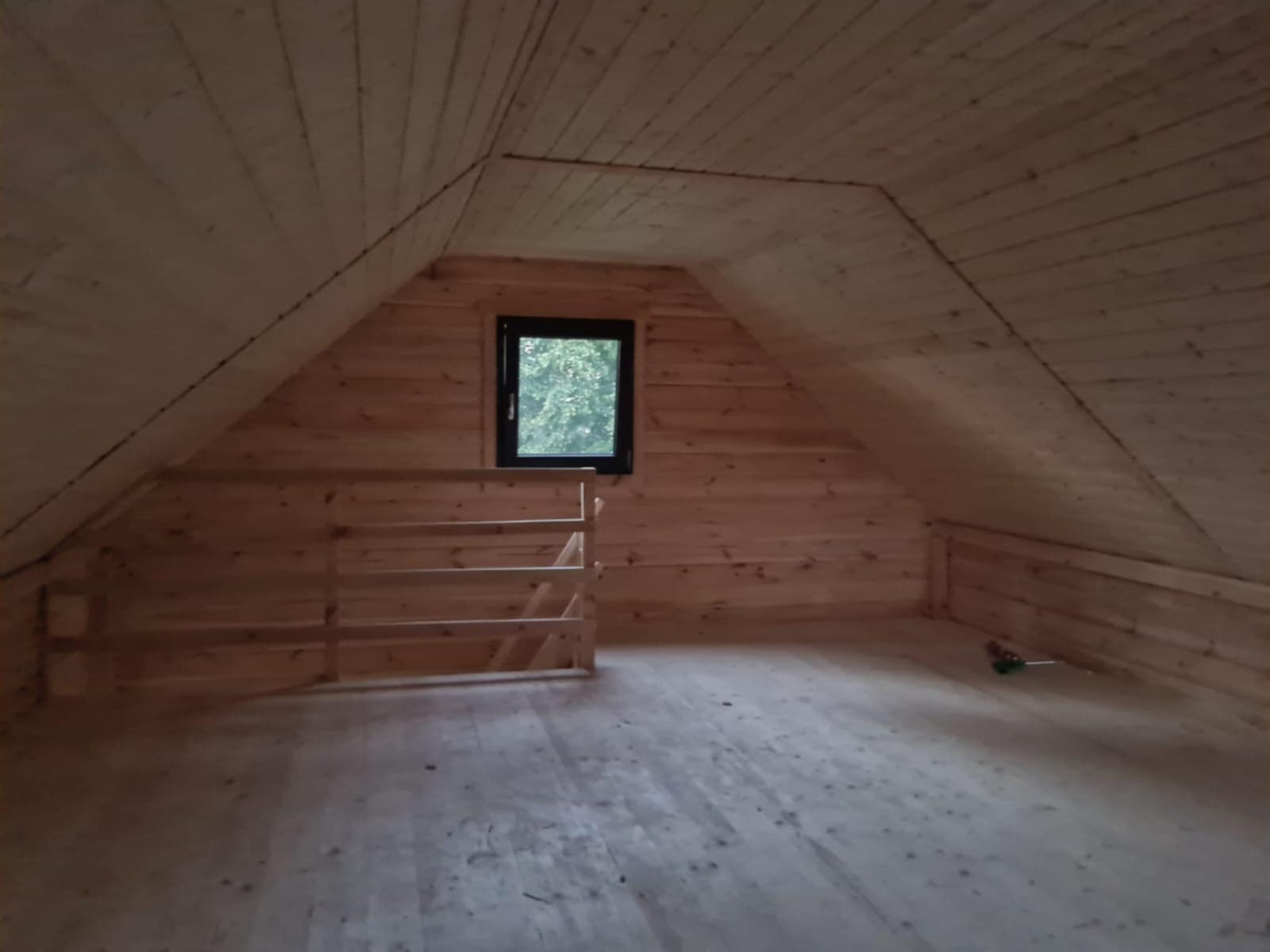 Surowe wnętrze poddasza domu z bali z oknem i drewnianą barierką. Widoczne deski na ścianach, podłodze i suficie. Minimalistyczny, naturalny wygląd.