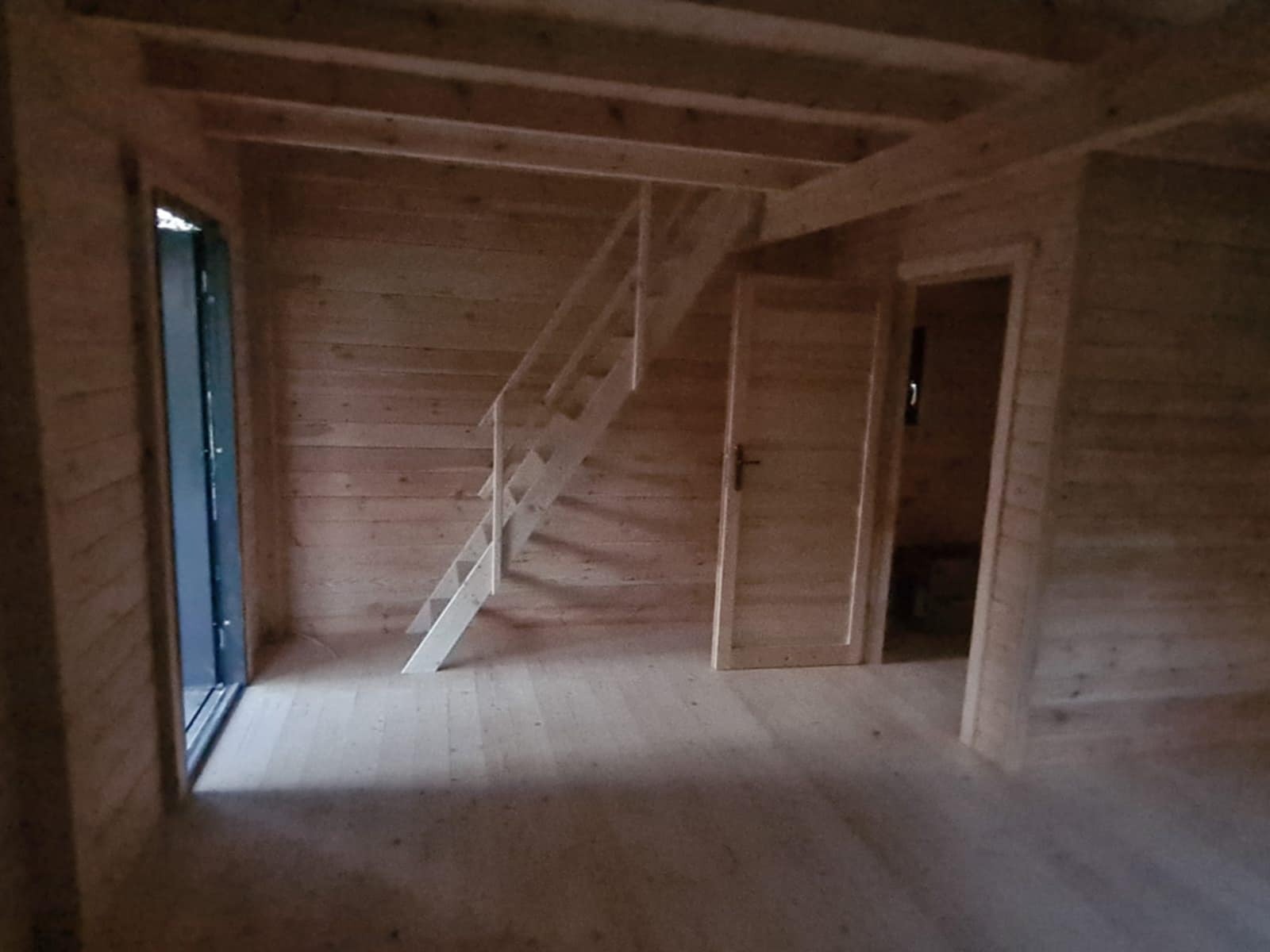 Wnętrze drewnianego domu w trakcie budowy: surowe ściany z bali, schody na antresolę i otwór drzwiowy. Widoczne naturalne usłojenie drewna.