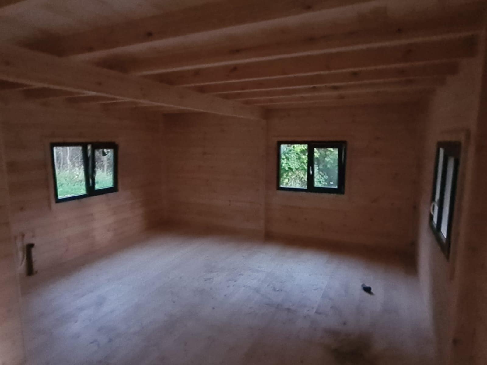 Wnętrze surowego drewnianego domu z trzema oknami w czarnych ramach. Widoczne drewniane ściany, podłoga i sufit z belek. Etap budowy domu z bali lub szkieletowego.