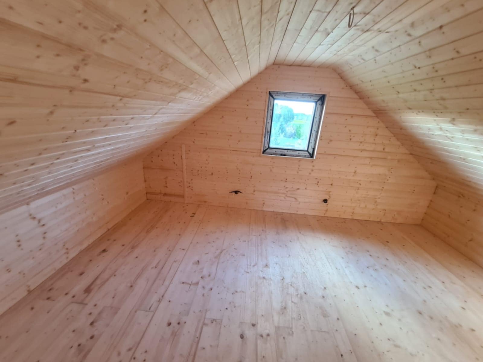 Drewniane wnętrze poddasza z oknem dachowym. Widoczne deski na podłodze, ścianach i suficie, tworzące ciepły, naturalny klimat. Surowe wykończenie.