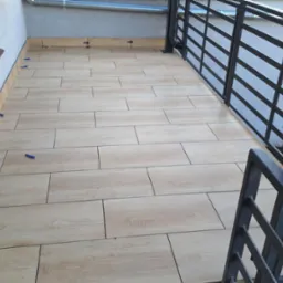 Balkon z ułożonymi płytkami imitującymi drewno, widoczne krzyżyki dystansowe. Metalowa balustrada w tle. Wykończone parapety.