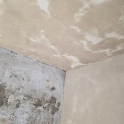 MIKO Erwin Mikołajczyk - Narożnik pokoju przed remontem: ściana z widocznymi plamami i ubytkami tynku, sufit z nierównomiernym wykończeniem, gotowy do dalszych prac wykończeniowych.