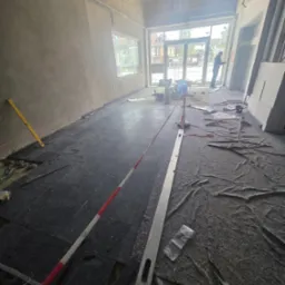 Wnętrze w trakcie układania ciemnej podłogi z płytek. Widoczne narzędzia, poziomica i folia ochronna na podłodze. W tle widoczne okno i osoba pracująca na zewnątrz.