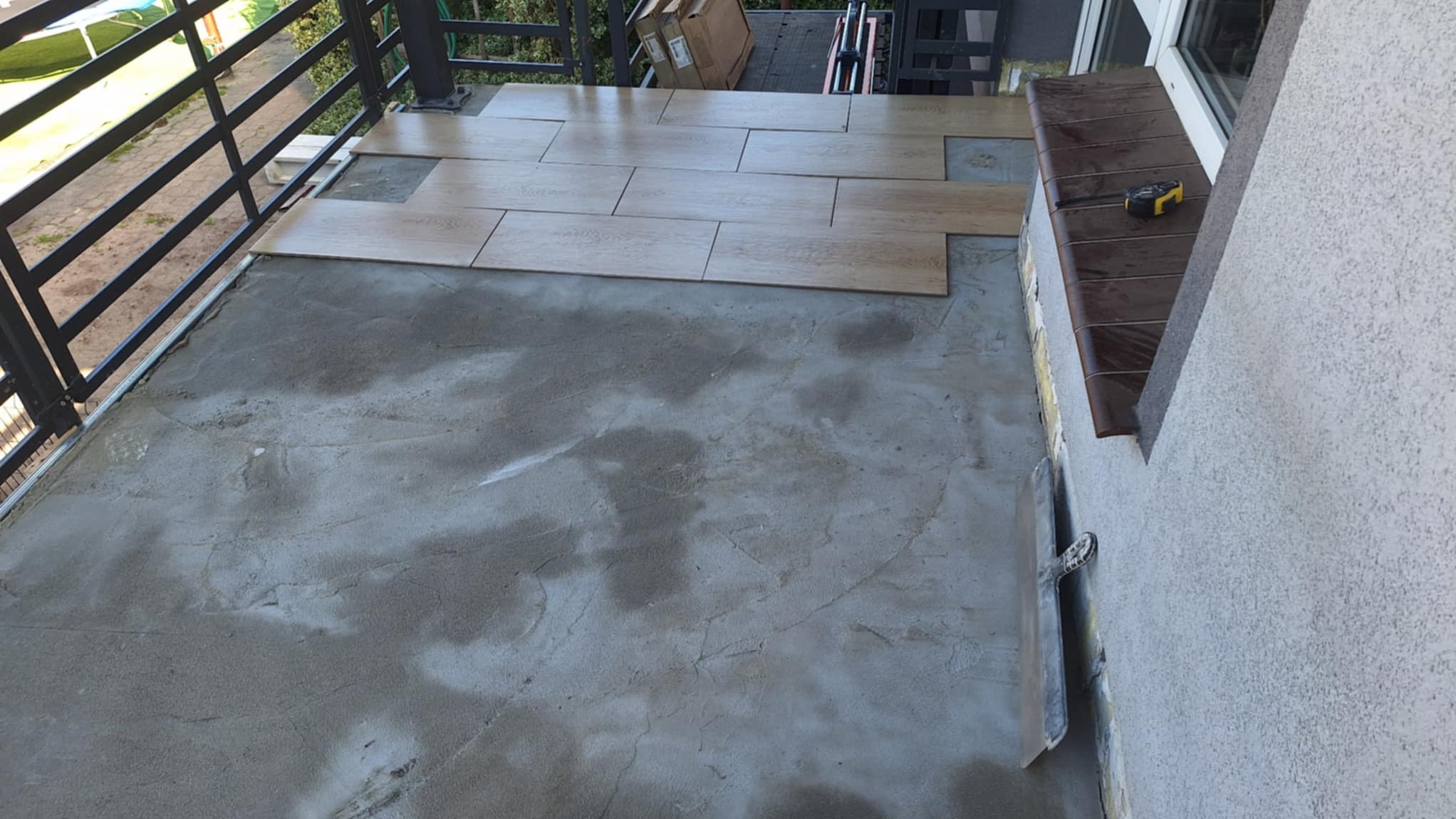 Balkon w trakcie układania płytek imitujących drewno. Część płytek już ułożona, widoczna zaprawa, poziomica. Balustrada balkonowa czarna, tło zielone.