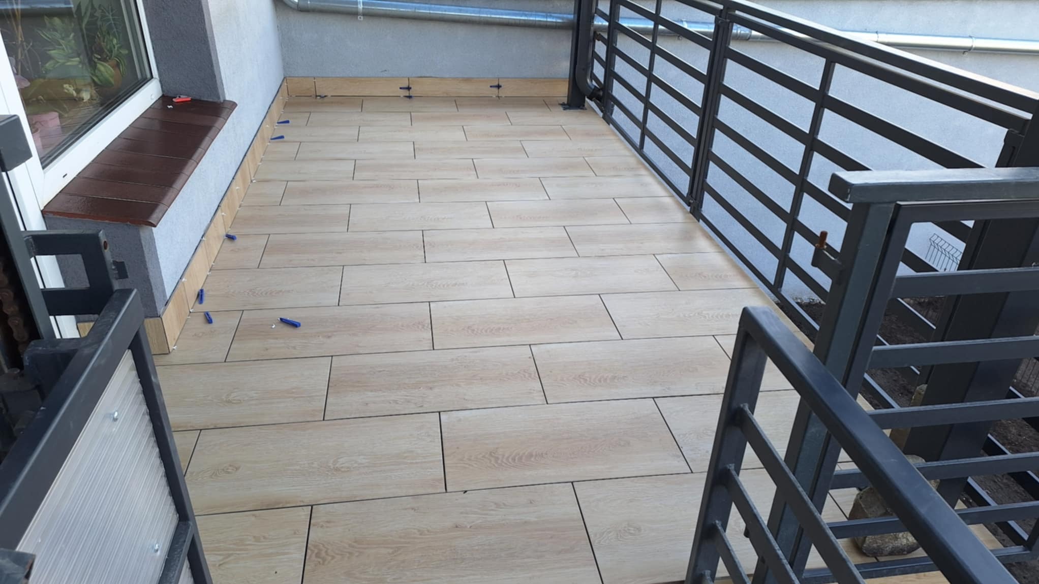 Balkon z ułożonymi płytkami imitującymi drewno, widoczne krzyżyki dystansowe. Metalowa balustrada w tle. Wykończone parapety.
