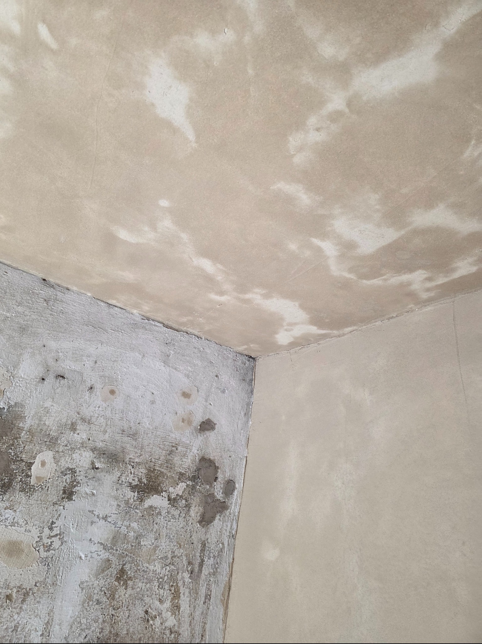 Narożnik pokoju przed remontem: ściana z widocznymi plamami i ubytkami tynku, sufit z nierównomiernym wykończeniem, gotowy do dalszych prac wykończeniowych.