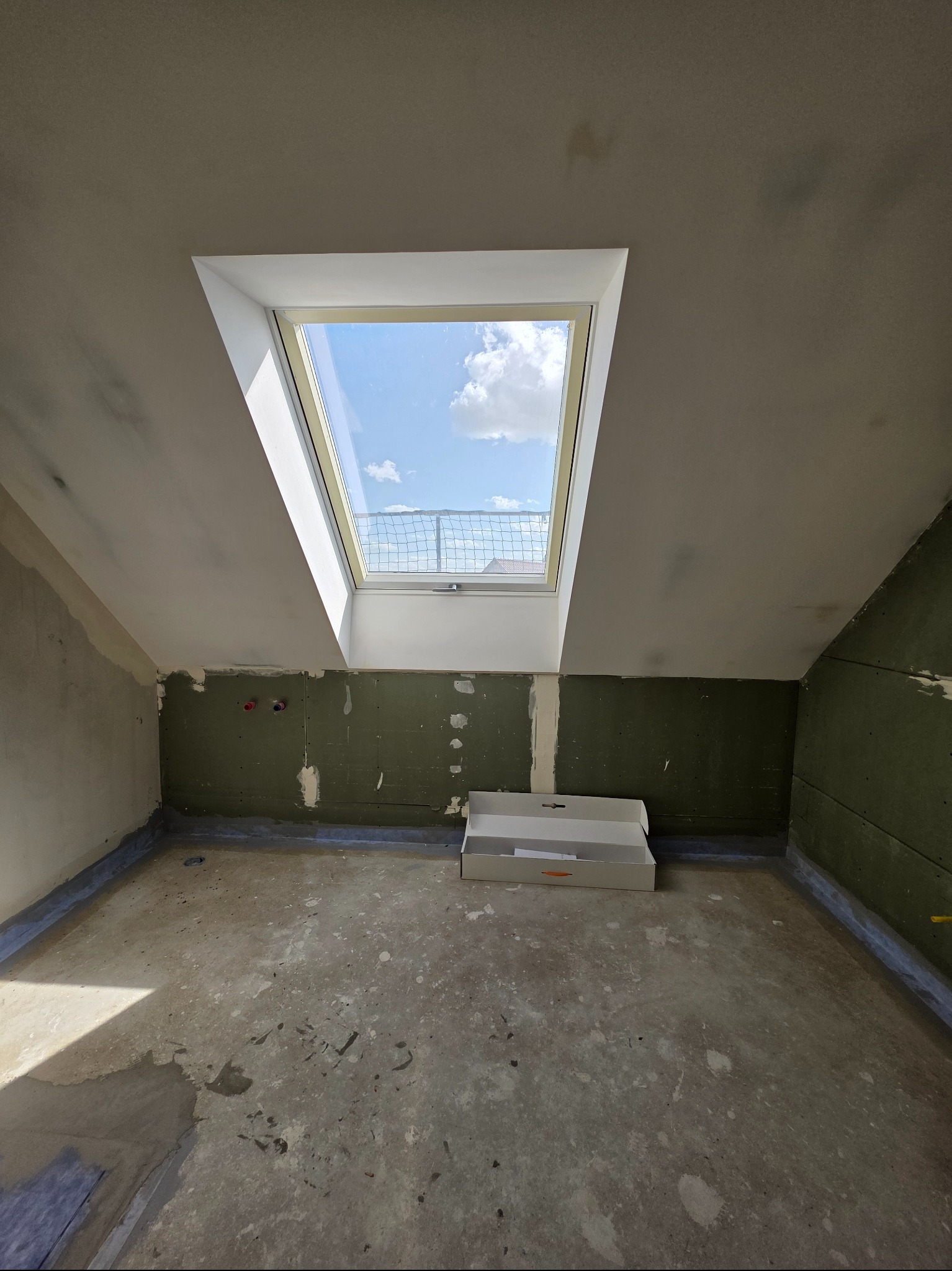 Poddasze w trakcie prac wykończeniowych: okno dachowe z widokiem na niebo, surowe ściany, przygotowane do dalszych prac. Otwarty karton na podłodze, widoczne ślady prac.