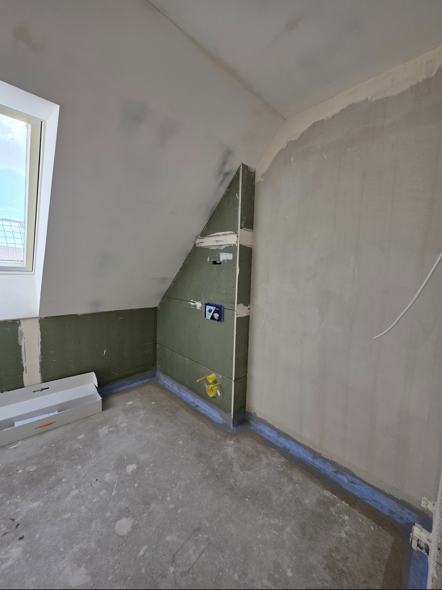 Poddasze w trakcie prac wykończeniowych: ściany z płyt gipsowych, fragment okna, przygotowane podłoże. Widoczne instalacje wodne i elektryczne.