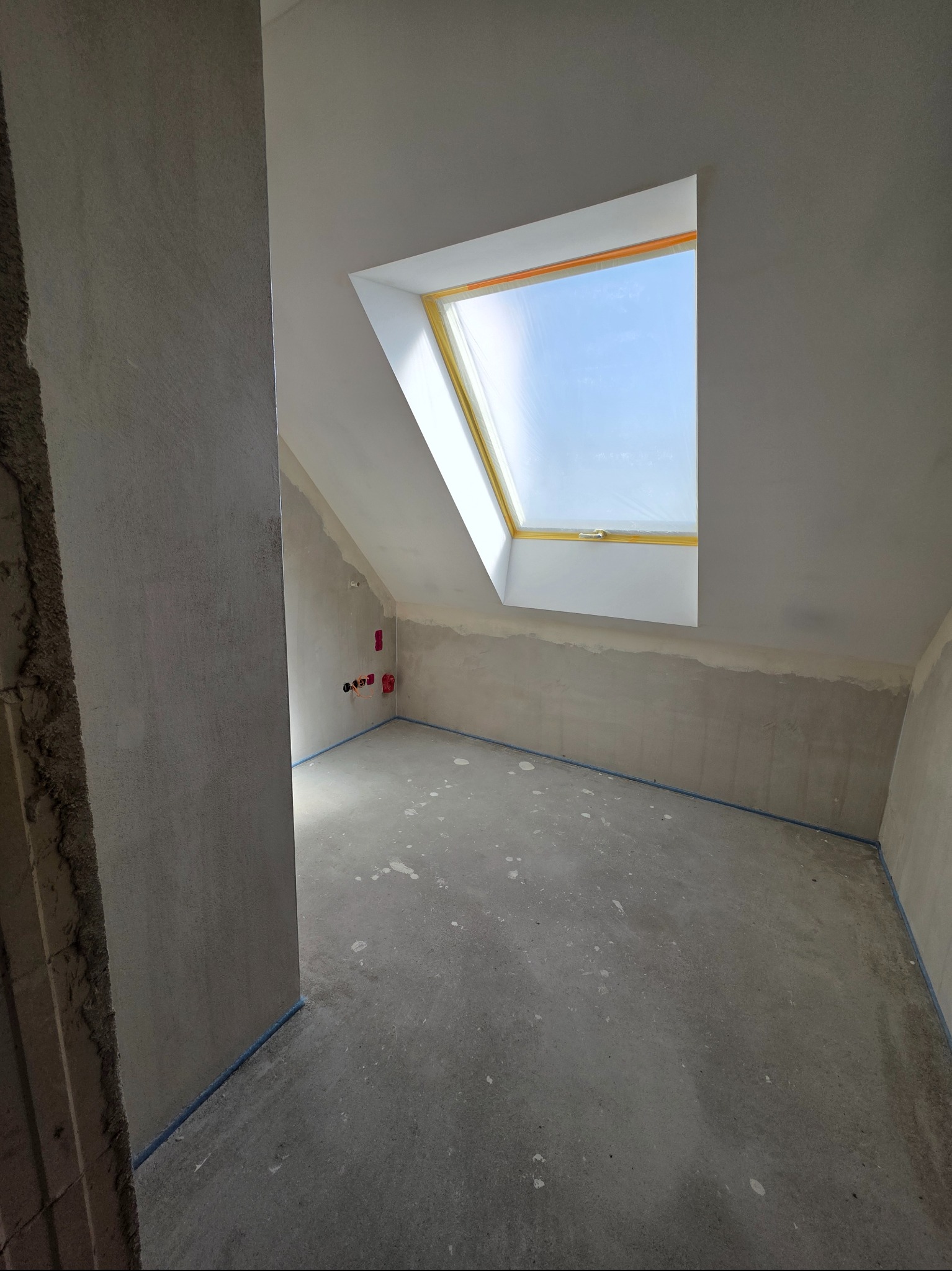 Surowe wnętrze poddasza z oknem dachowym i instalacją elektryczną. Ściany przygotowane do malowania, podłoga betonowa z widocznymi śladami zaprawy.
