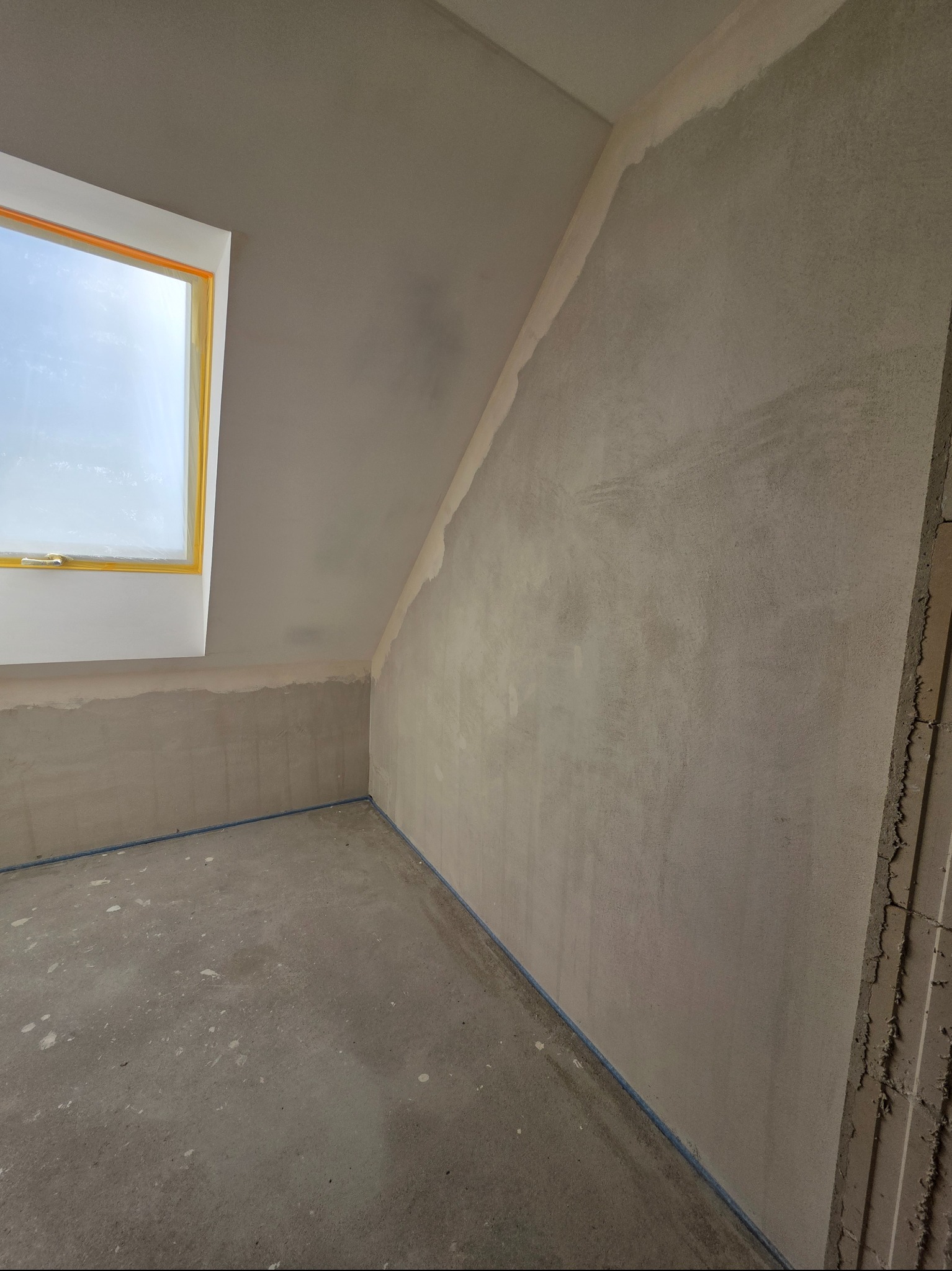 Pokój na poddaszu w trakcie prac wykończeniowych. Ściany pokryte gładzią, okno dachowe z żółtą ramą, podłoga zabezpieczona folią. Widoczne ślady po pracach budowlanych.