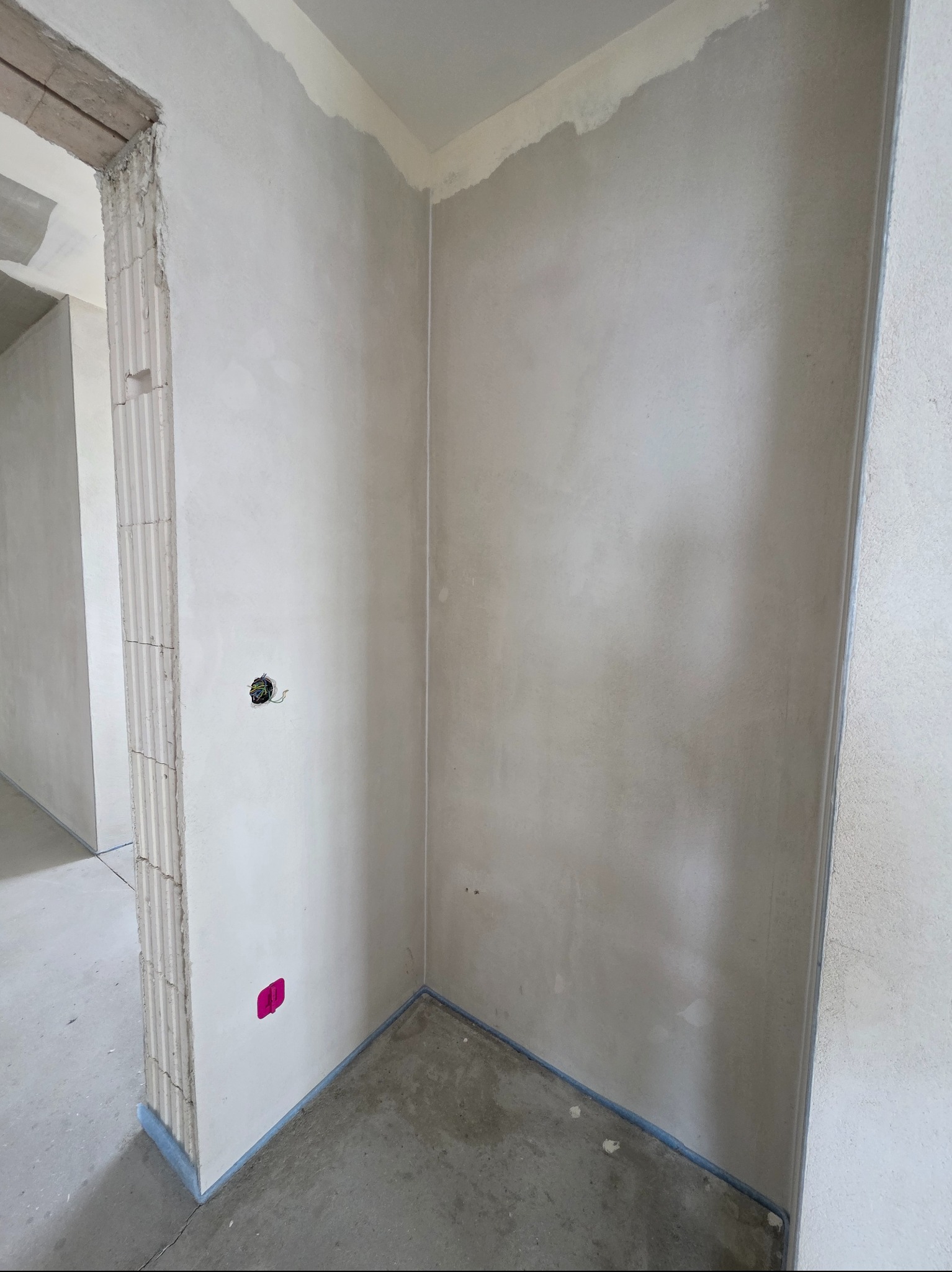 Surowe ściany w rogu pokoju po nałożeniu gładzi szpachlowej, zabezpieczone folią malarską przy podłodze i futrynie. Widoczne kable w puszce elektrycznej.