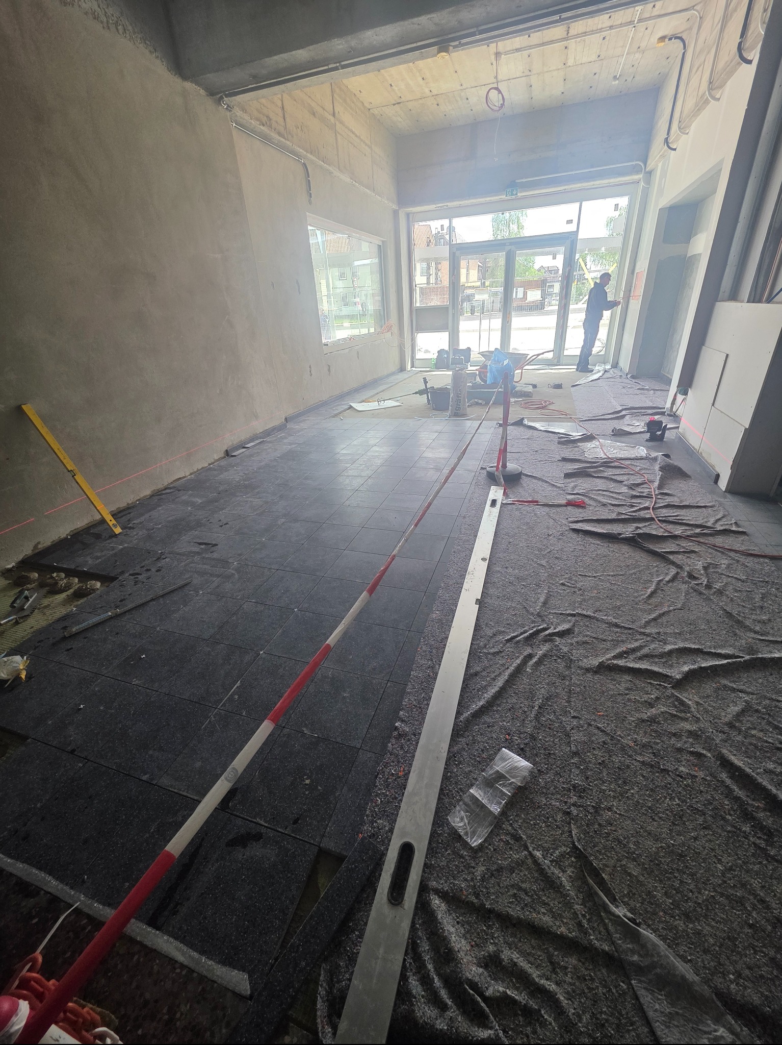 Wnętrze w trakcie układania ciemnej podłogi z płytek. Widoczne narzędzia, poziomica i folia ochronna na podłodze. W tle widoczne okno i osoba pracująca na zewnątrz.