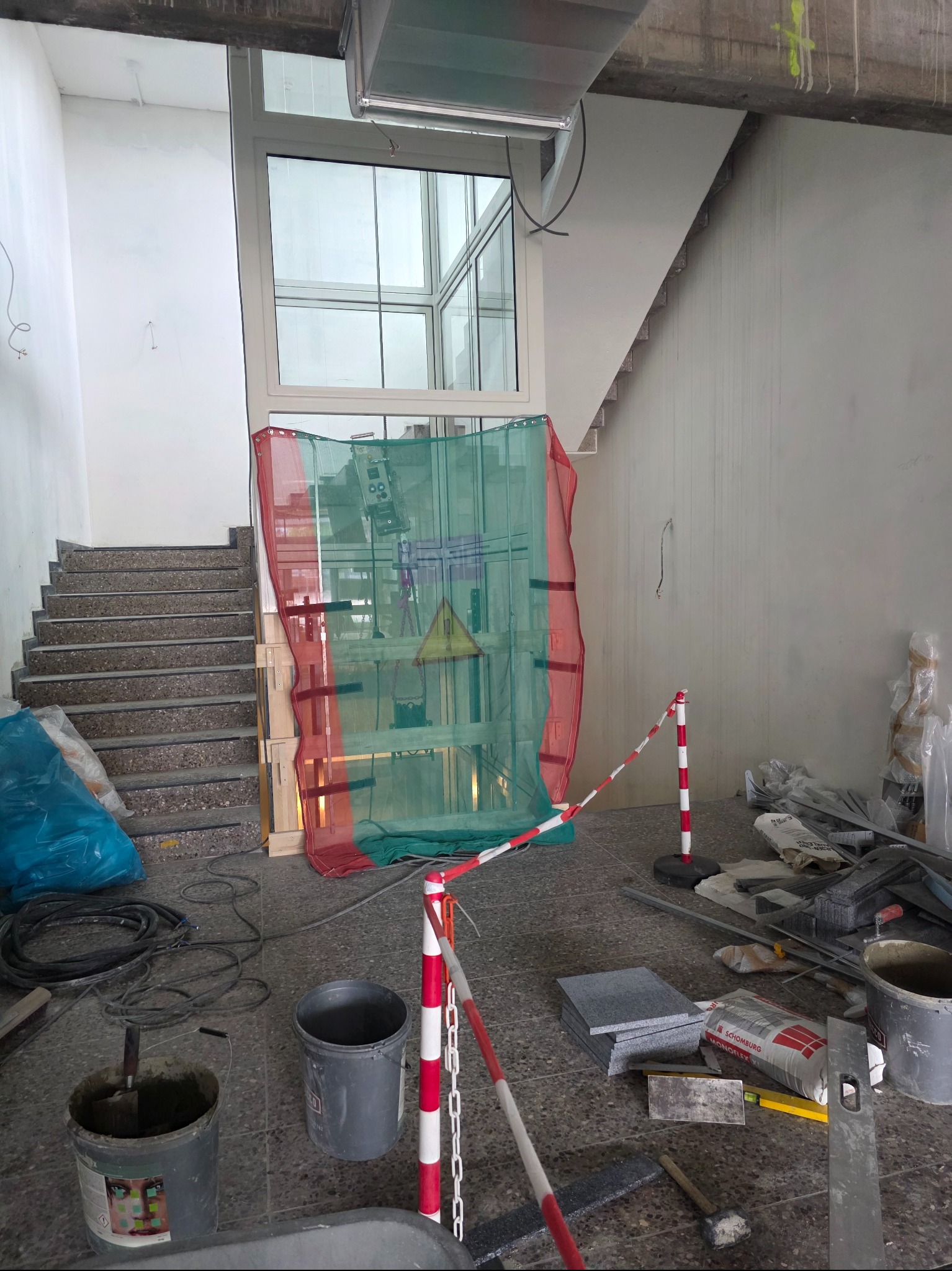 Wnętrze w trakcie prac wykończeniowych: widoczne schody, okno, materiały budowlane, wiadro z zaprawą i zabezpieczenia. Etap remontu w surowym stanie.