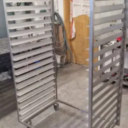 W&oacute;zek transportowy ze stali nierdzewnej z p&oacute;łkami szczelinowymi na k&oacute;łkach, widok z przodu, w tle ściana i drzwi z instrukcją BHP.