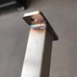 Szlifowany profil stalowy z przyspawaną płytką z otworem. Widoczne ślady spawania TIG z charakterystycznymi kolorami. Detal konstrukcyjny.