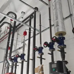Instalacja przemysłowa z rurami, zaworami i izolacją, zamontowana przy białej, teksturowanej ścianie. Widoczne fragmenty konstrukcji wsporczej.