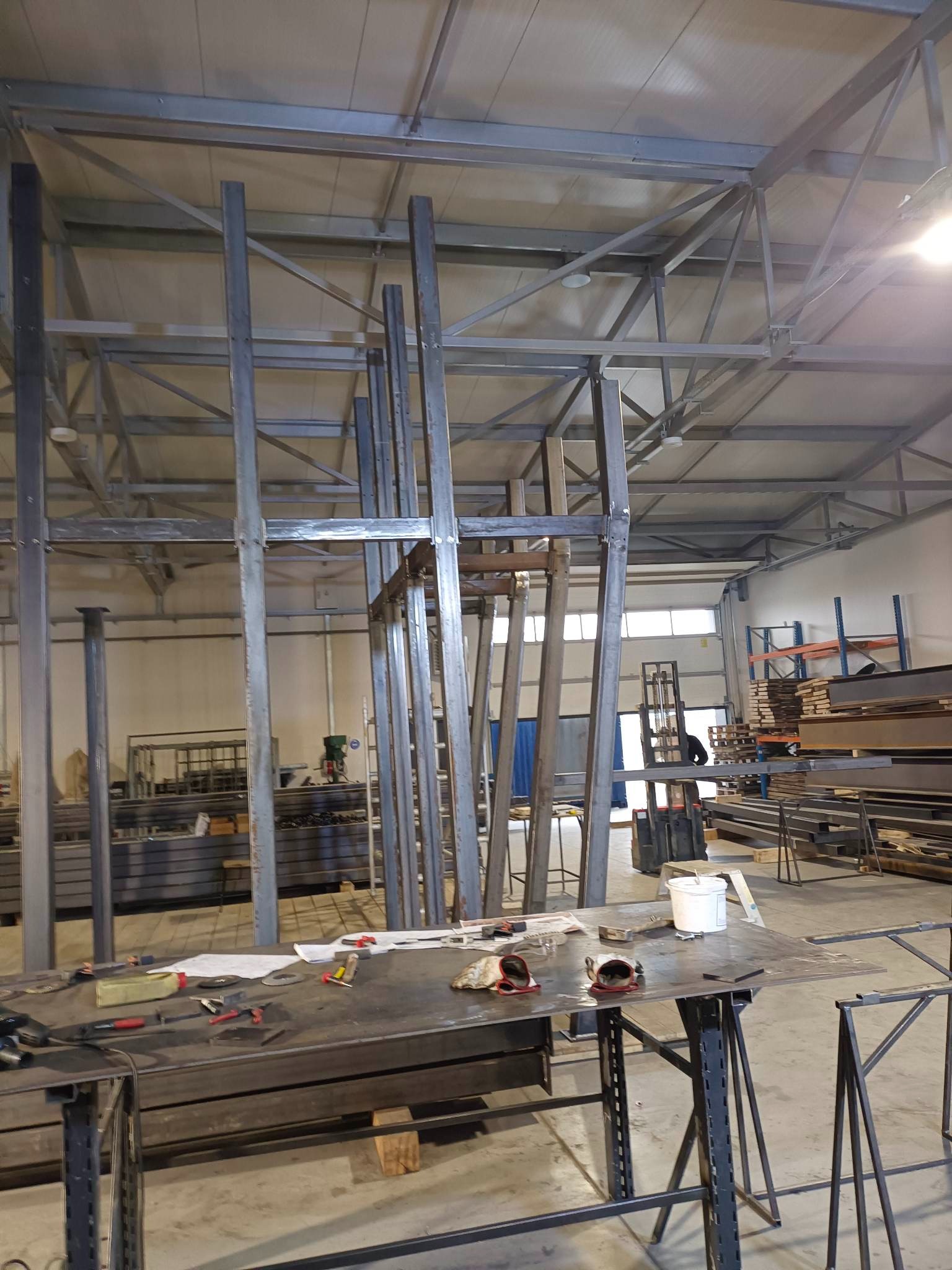 Wnętrze hali produkcyjnej z konstrukcją stalową w trakcie montażu. Widoczne narzędzia na stole warsztatowym i elementy konstrukcyjne. Charakterystyczne oświetlenie hali.