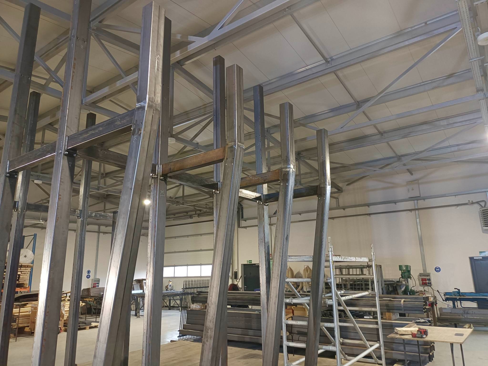 Konstrukcja metalowa w hali produkcyjnej, spawane elementy stalowe, widoczne spoiny, surowy stan, drabina, oświetlenie przemysłowe, przygotowanie do montażu.