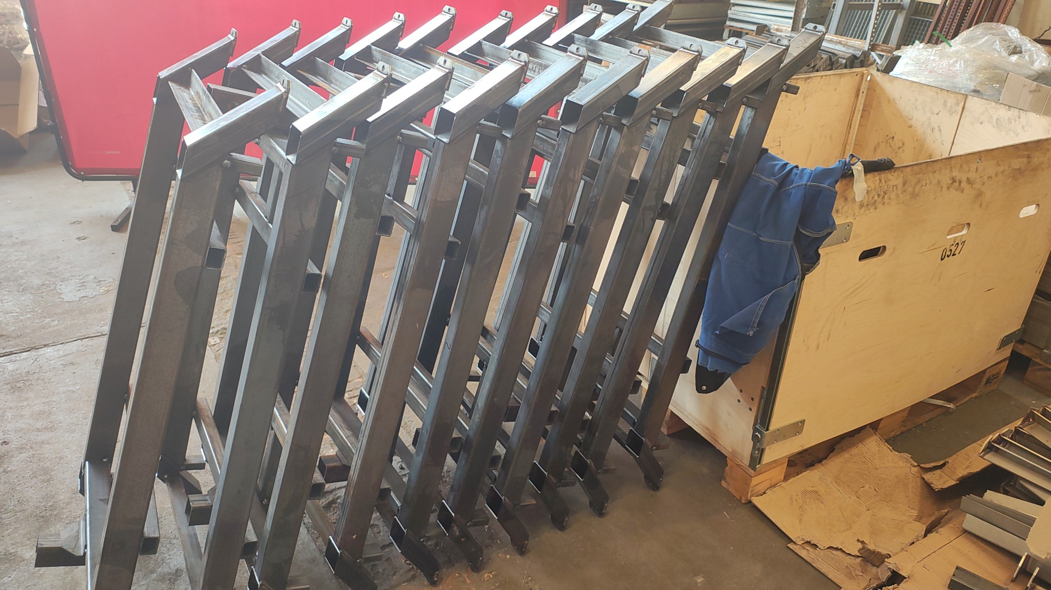 Stos metalowych konstrukcji ramowych, prawdopodobnie elementów balustrad lub ogrodzeń, opartych o drewnianą skrzynię w warsztacie. Widoczny strój roboczy.