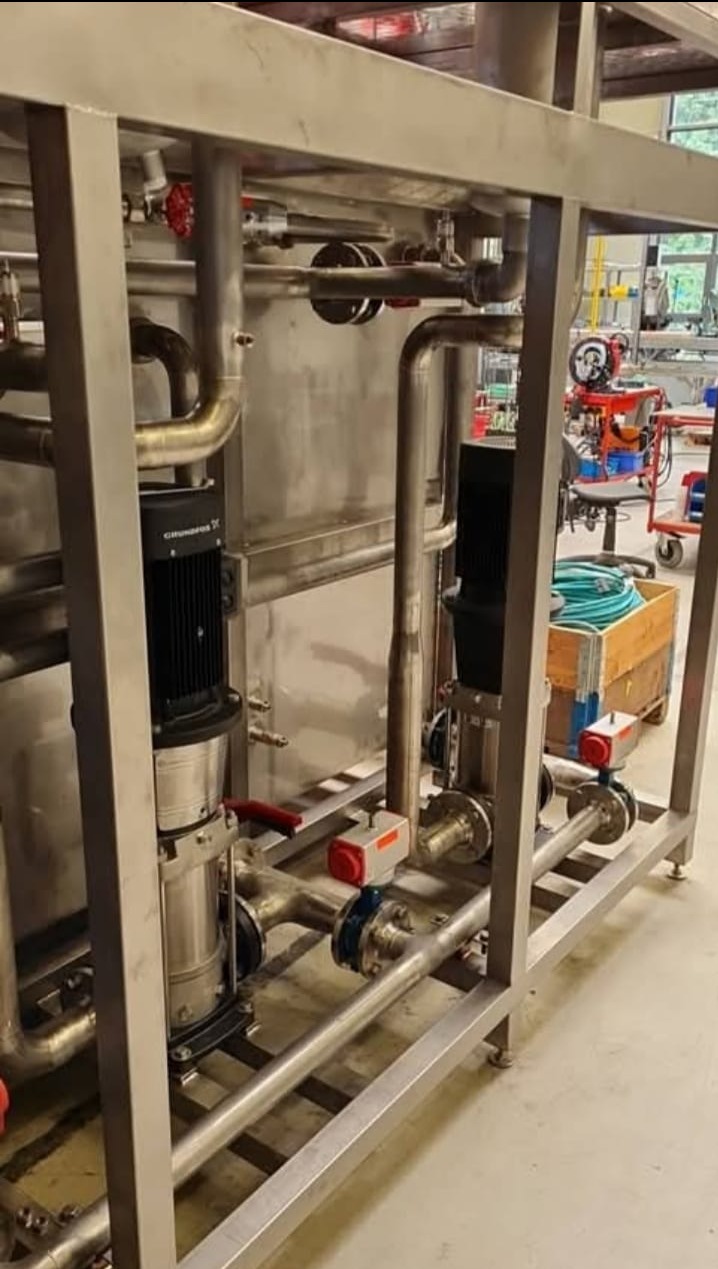 Kompleksowa instalacja przemysłowa ze stali nierdzewnej: rury, pompy Grundfos i zawory, osadzona w ramie z profili. Widoczne elementy spawane i skręcane.